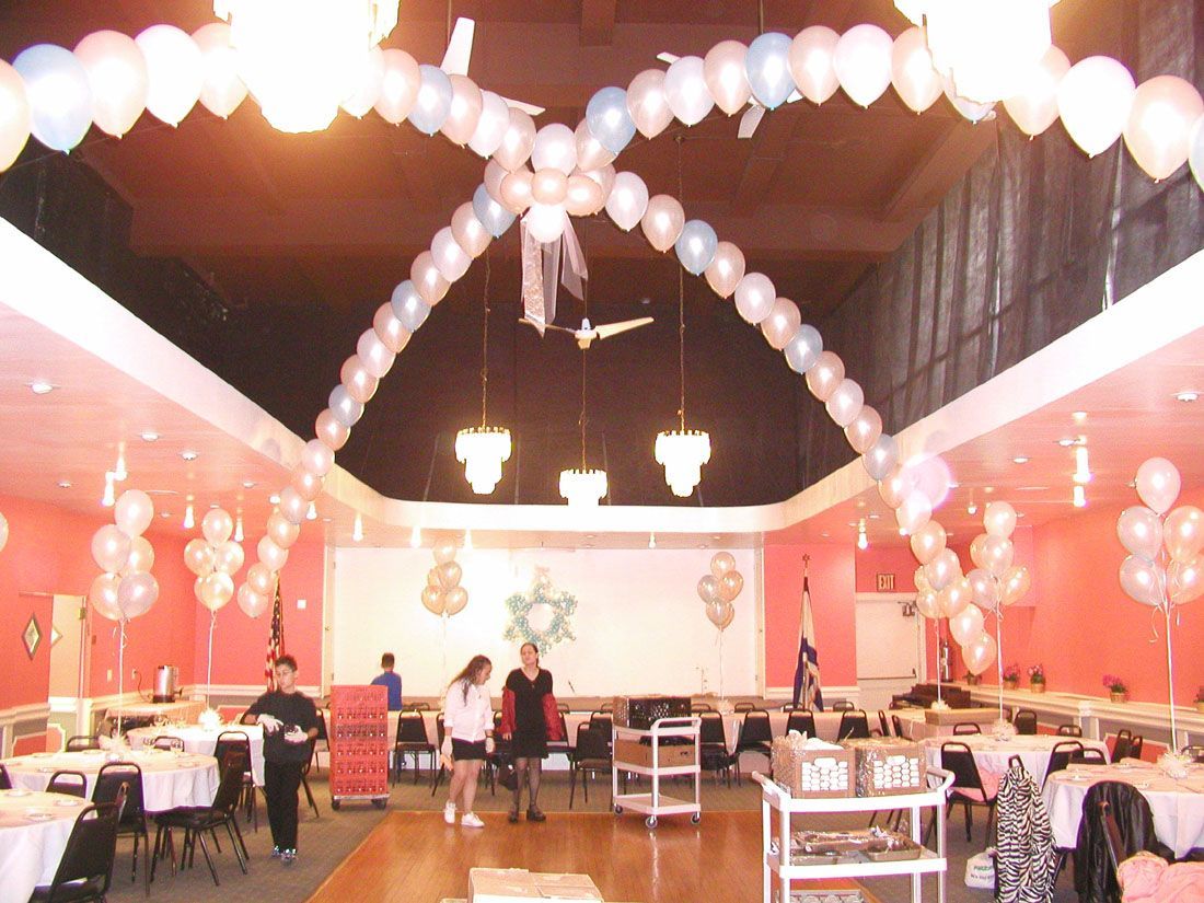 A decorated event hall with balloon arches and clusters. People are setting up tables.