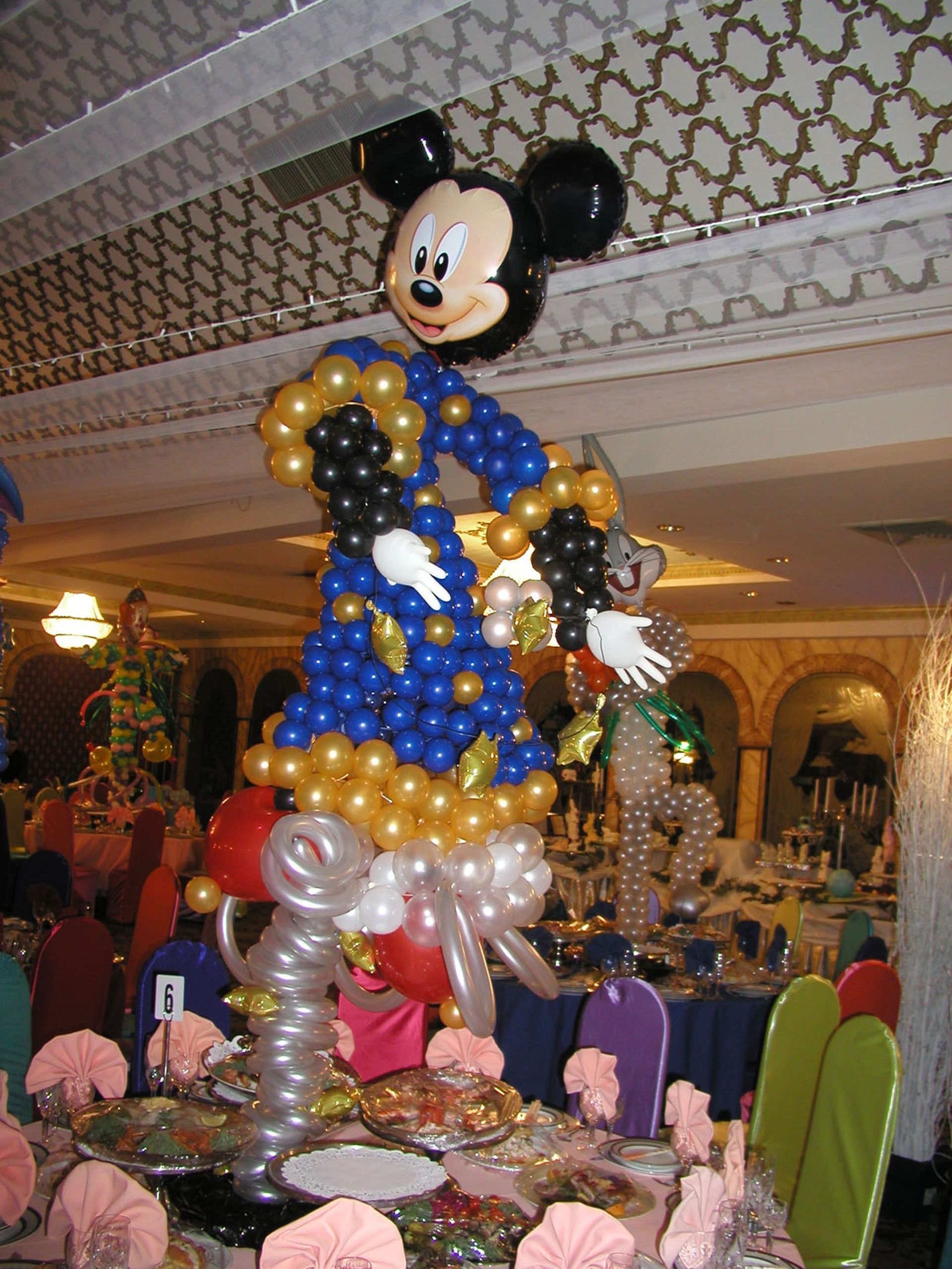 Balloon sculpture of Mickey Mouse in a blue wizard outfit, atop a decorated table in a banquet hall.