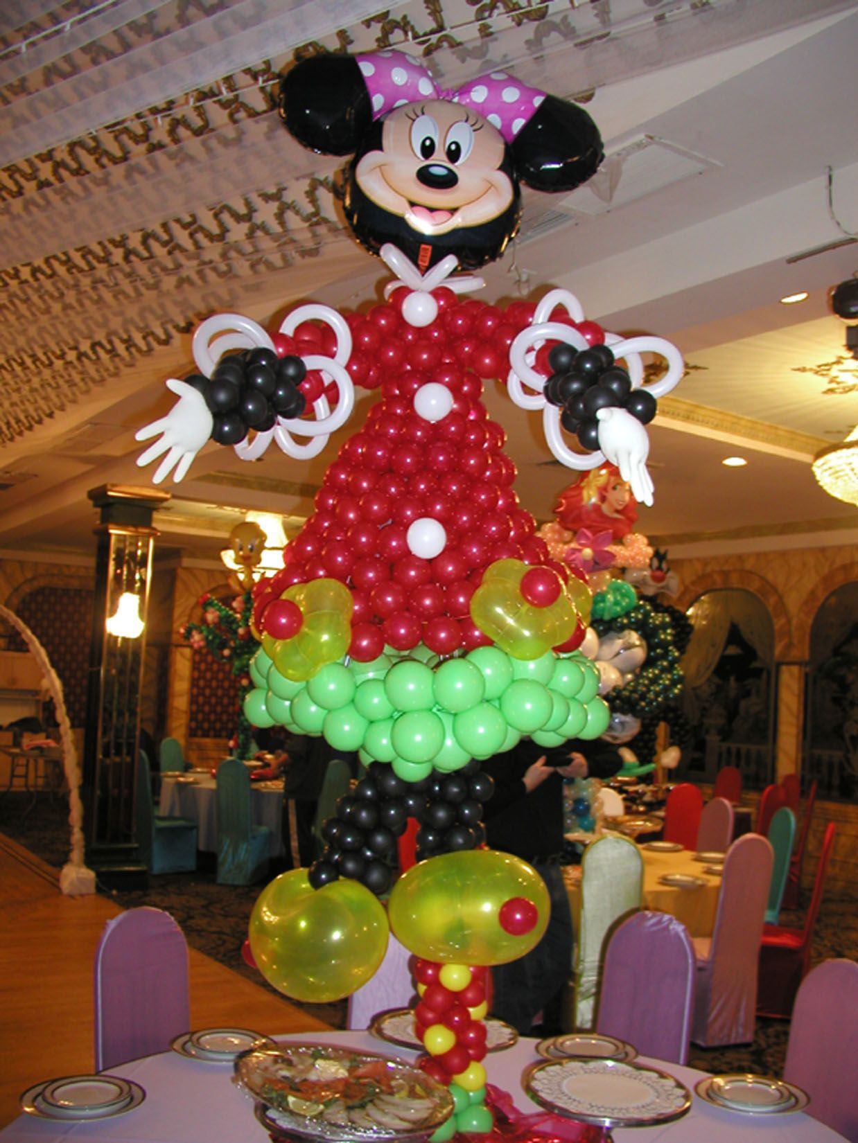 Balloon sculpture of Minnie Mouse in a red dress with white buttons, standing on a table in a decorated room.