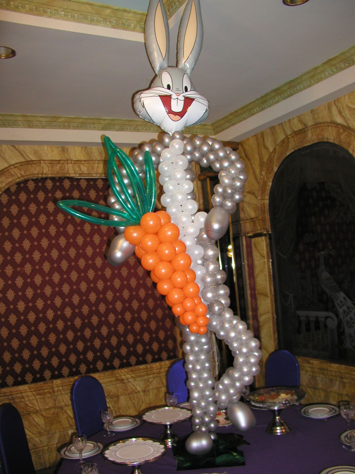 Bugs Bunny balloon sculpture holding a large orange carrot at a party table with purple tablecloth.