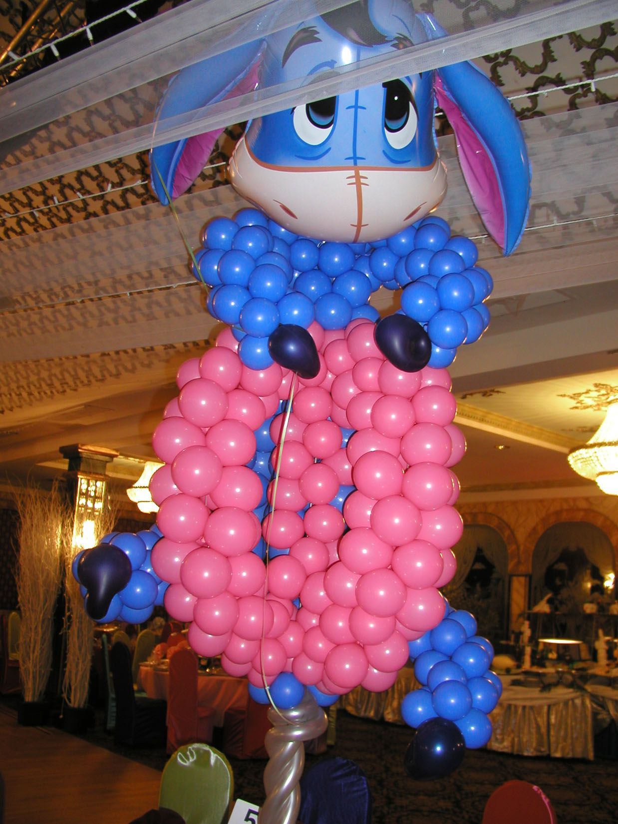 Balloon sculpture of Eeyore from Winnie the Pooh, blue and pink, standing in a decorated room.