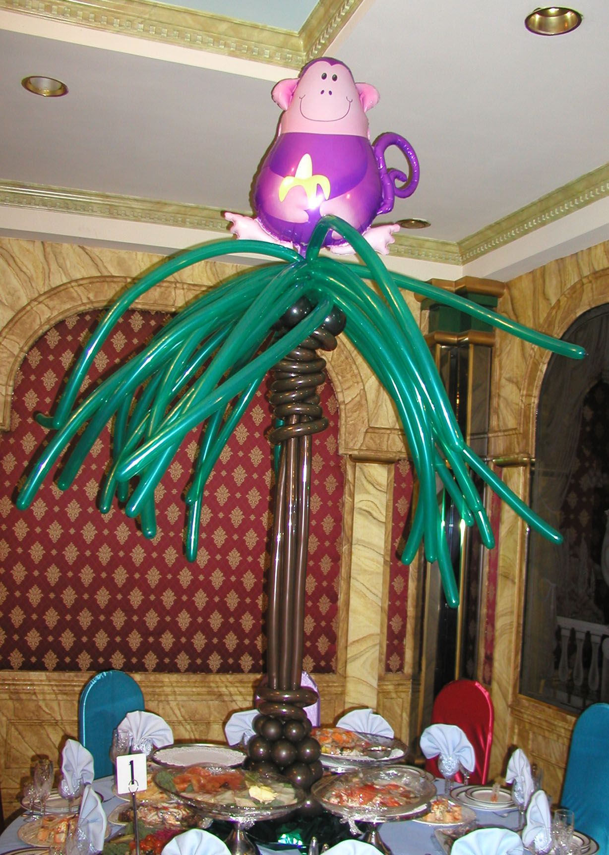 Balloon sculpture of a monkey in a teacup on a palm tree, green and brown balloons, set at a table.