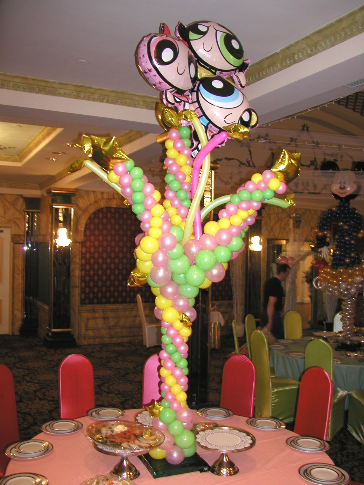 Balloon arrangement of Powerpuff Girls atop a table in a decorated banquet hall.