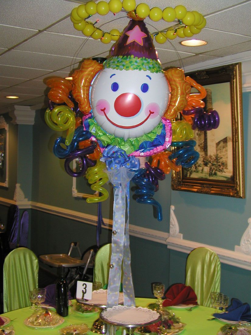 Clown balloon centerpiece with yellow, orange, and blue accents on a table with green tablecloth and chairs.