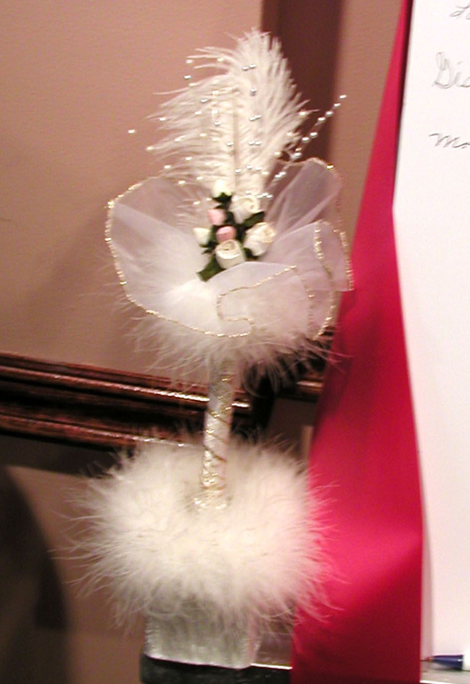 White feather pen decorated with tulle, pearls, and small flowers, next to a red ribbon.