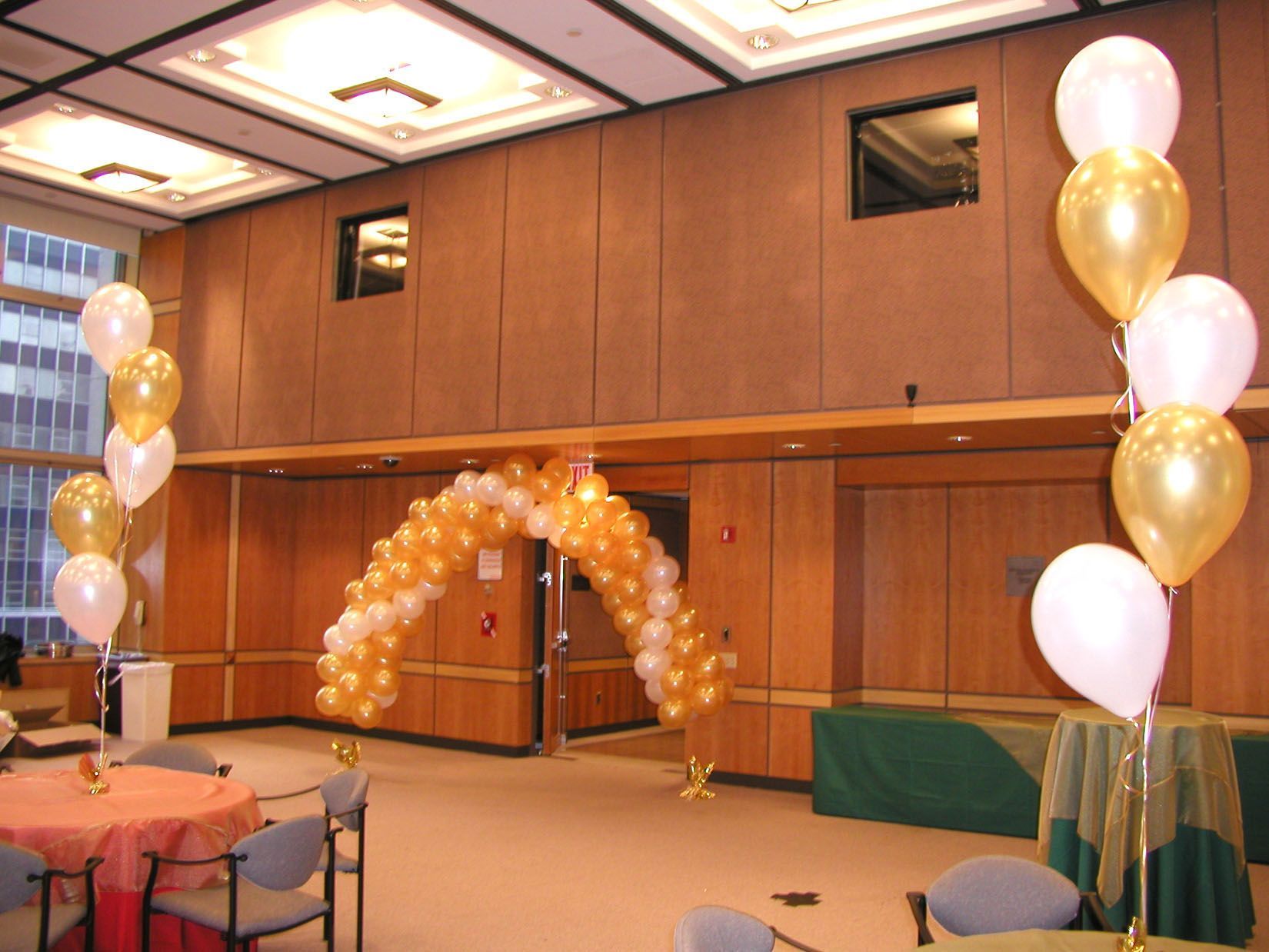 Balloon arch and columns in gold and white decorate a room with tables and chairs.