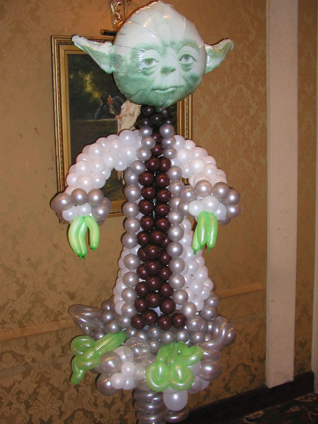 Balloon sculpture of Yoda, featuring green head and hands, white and gray body, brown neck.