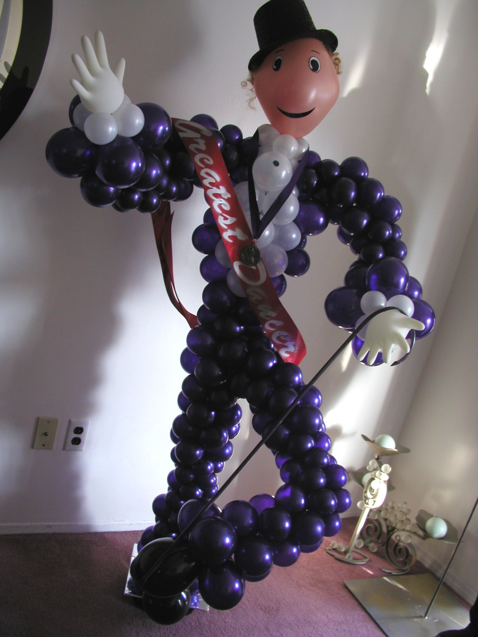 Balloon sculpture of a person in a purple tuxedo, waving. Face is a beige balloon, top hat.