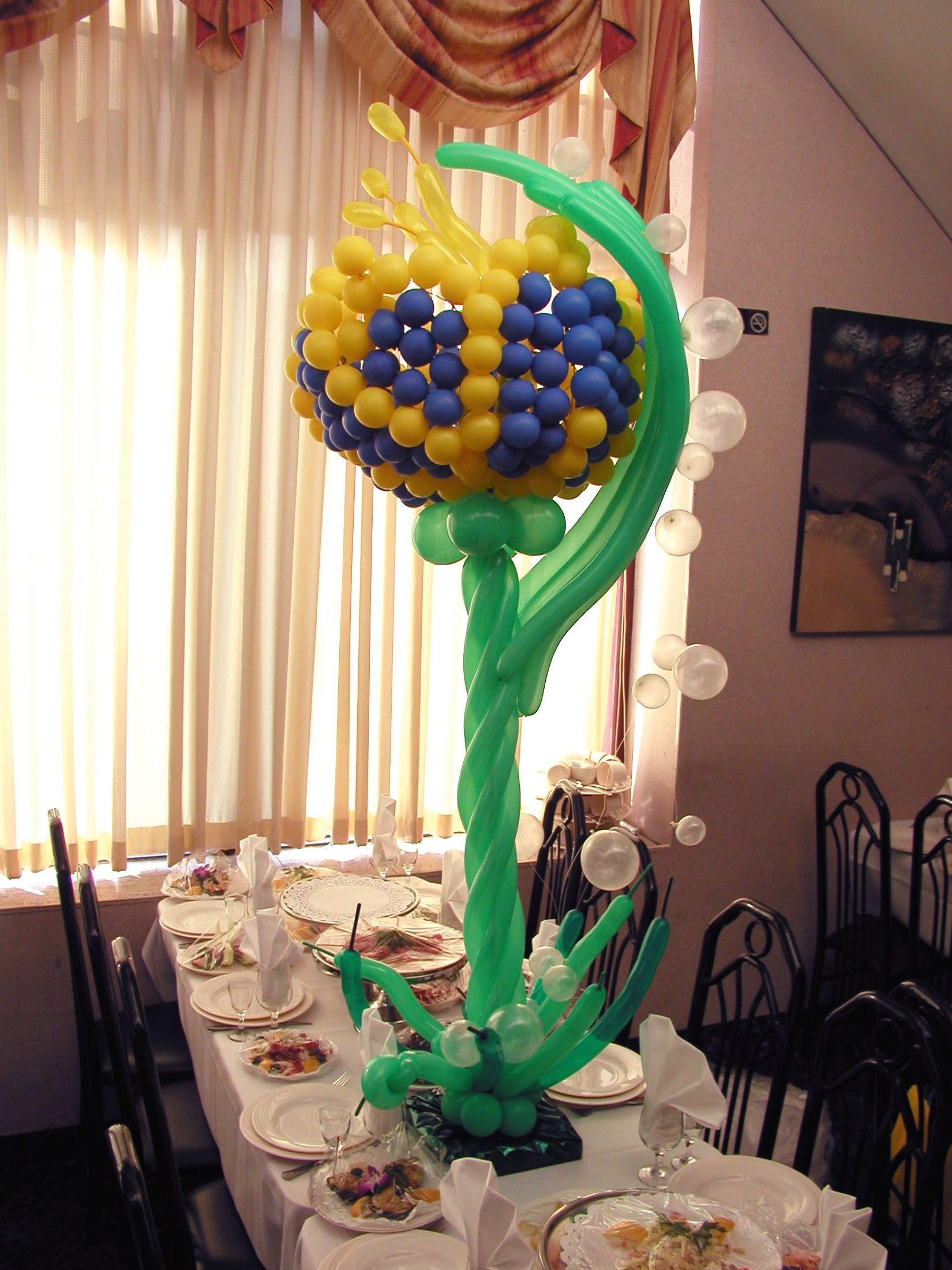Balloon sculpture centerpiece on a table, resembling a flower with yellow and blue bloom.