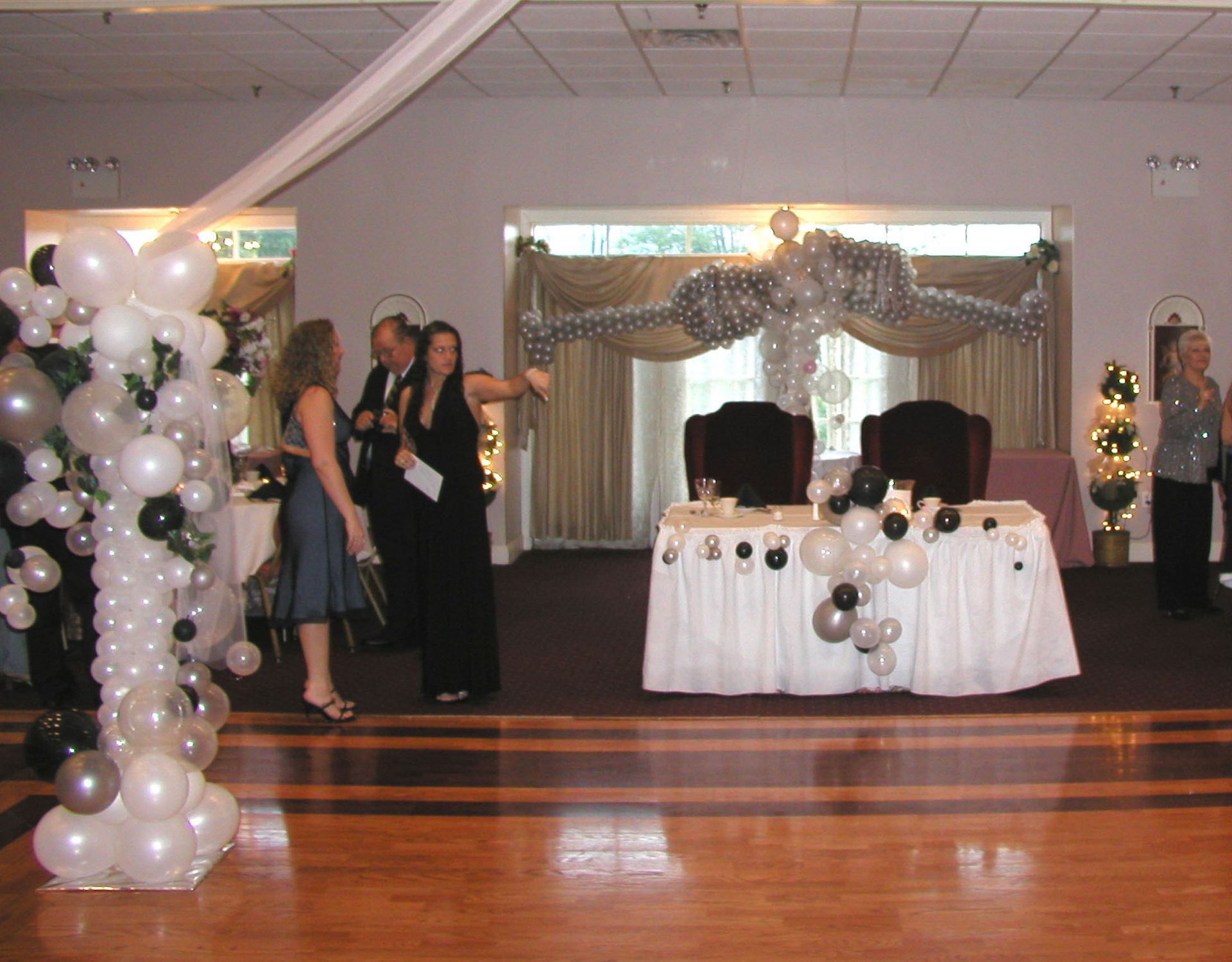 Formal event: people on a stage with balloon decorations. Two people stand near a decorated table, others on the sides.