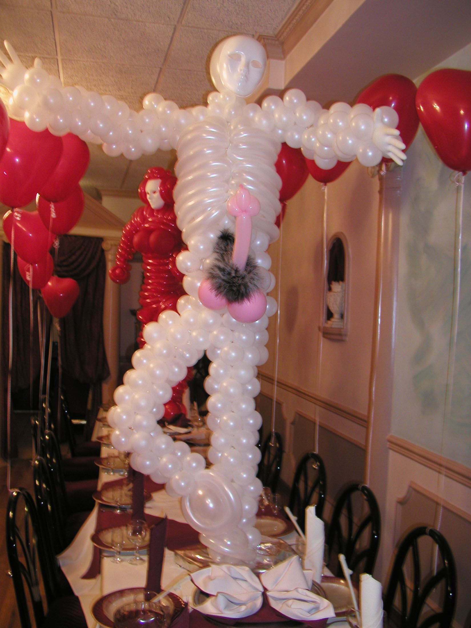 Balloon sculpture of a person with outstretched arms and genitalia, on a table. Red heart balloons and another balloon figure are in the background.