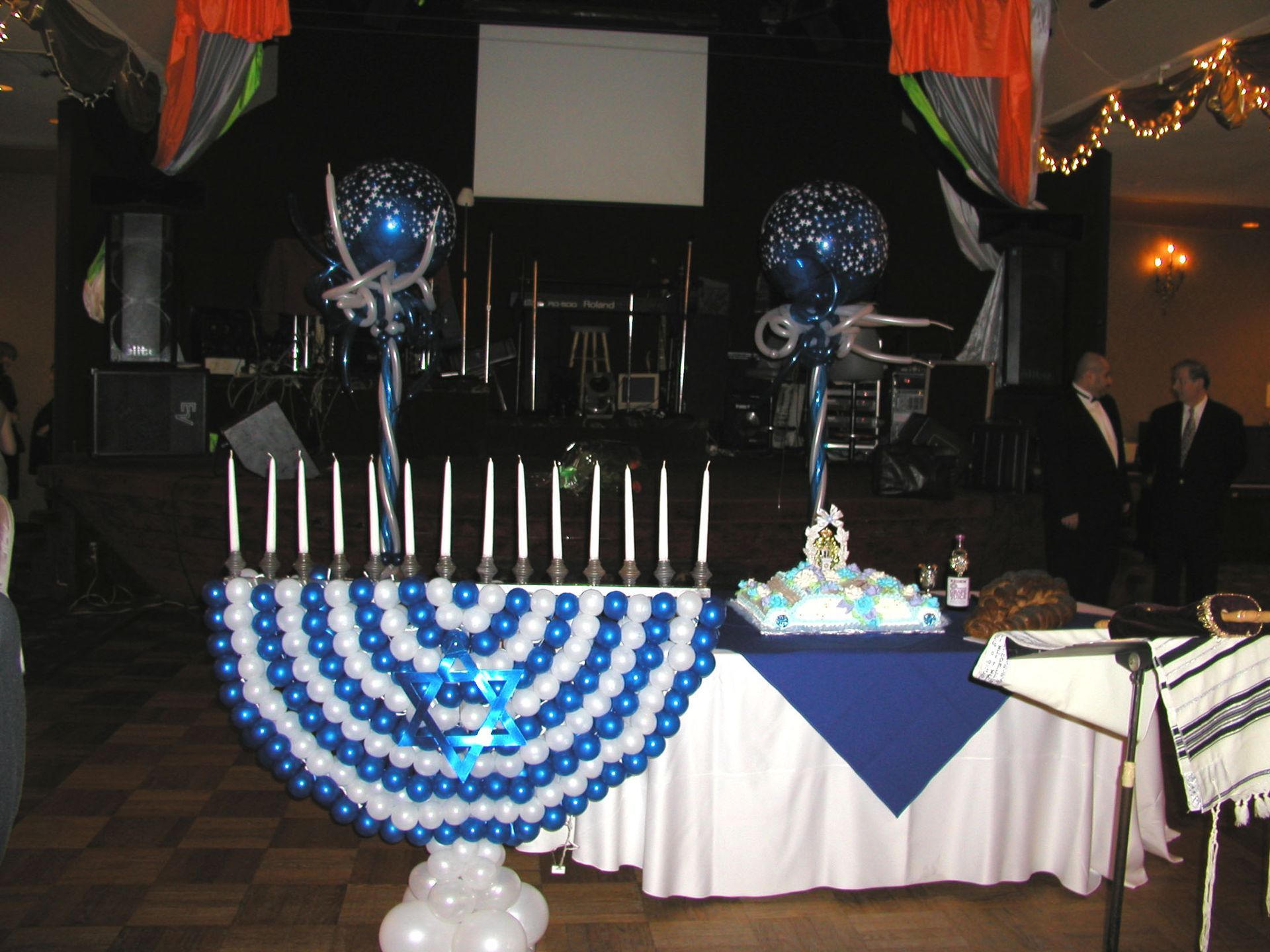 Balloon menorah and cake on a table at an event. A stage with a screen is in the background.