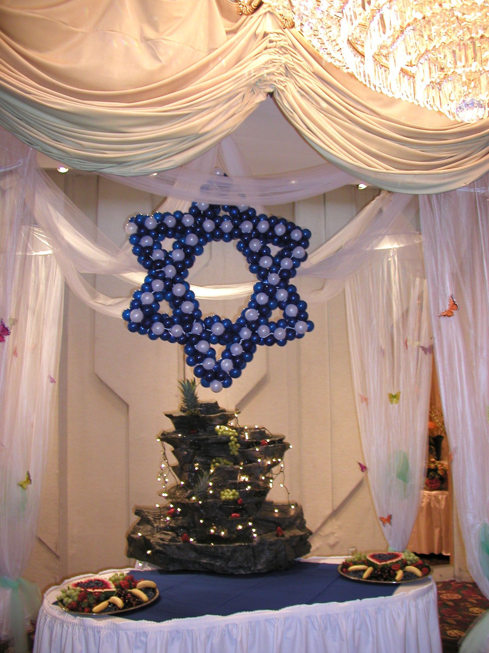Star of David made of blue and white balloons, above a decorated evergreen, draped with white fabric.