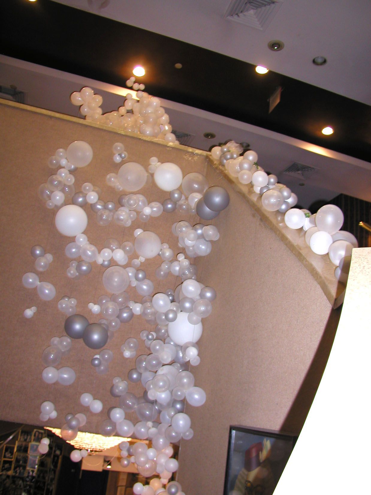 White and silver balloons cascading down a beige wall, possibly an indoor event decoration.