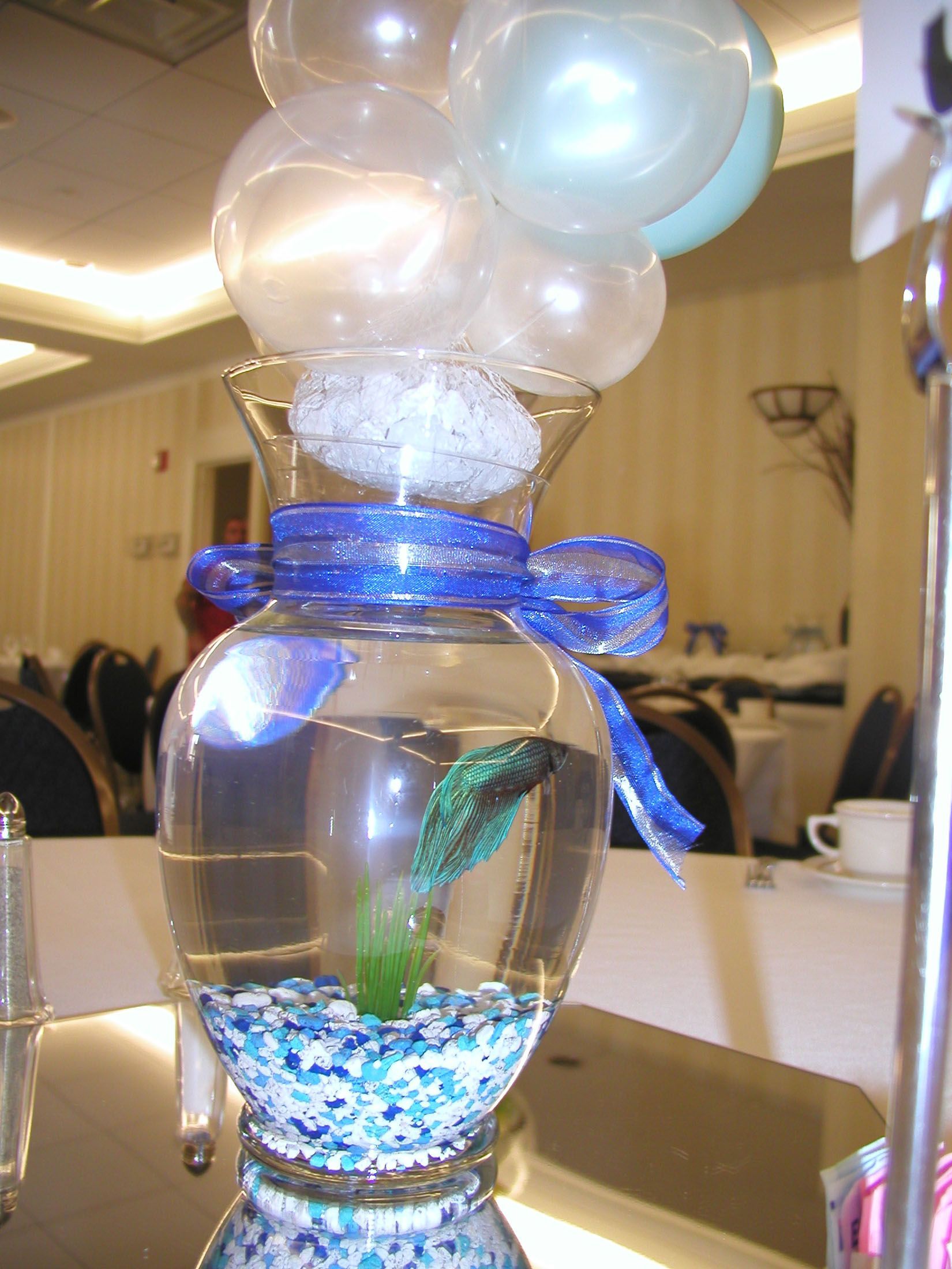 Fishbowl centerpiece with betta fish, blue ribbon, and balloons on a table.
