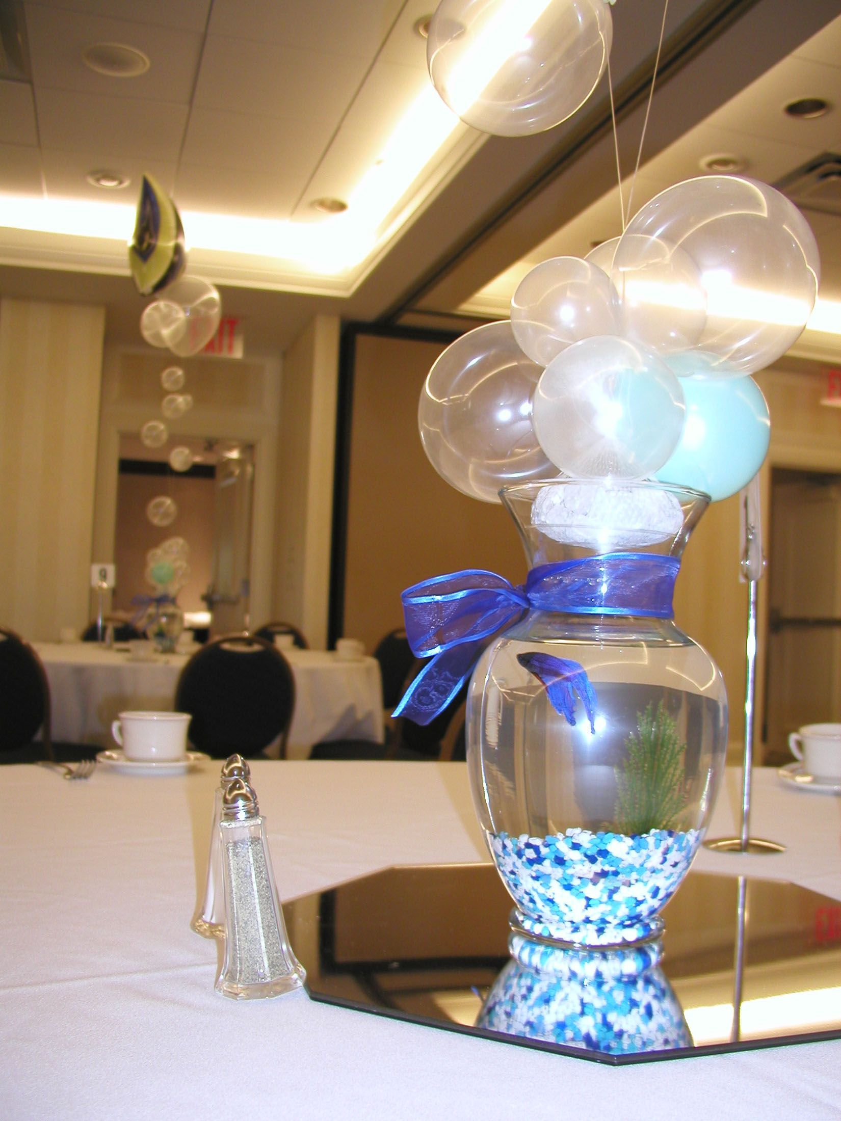 Centerpiece: Clear vase with blue ribbon, water, and balloons, on mirrored table.