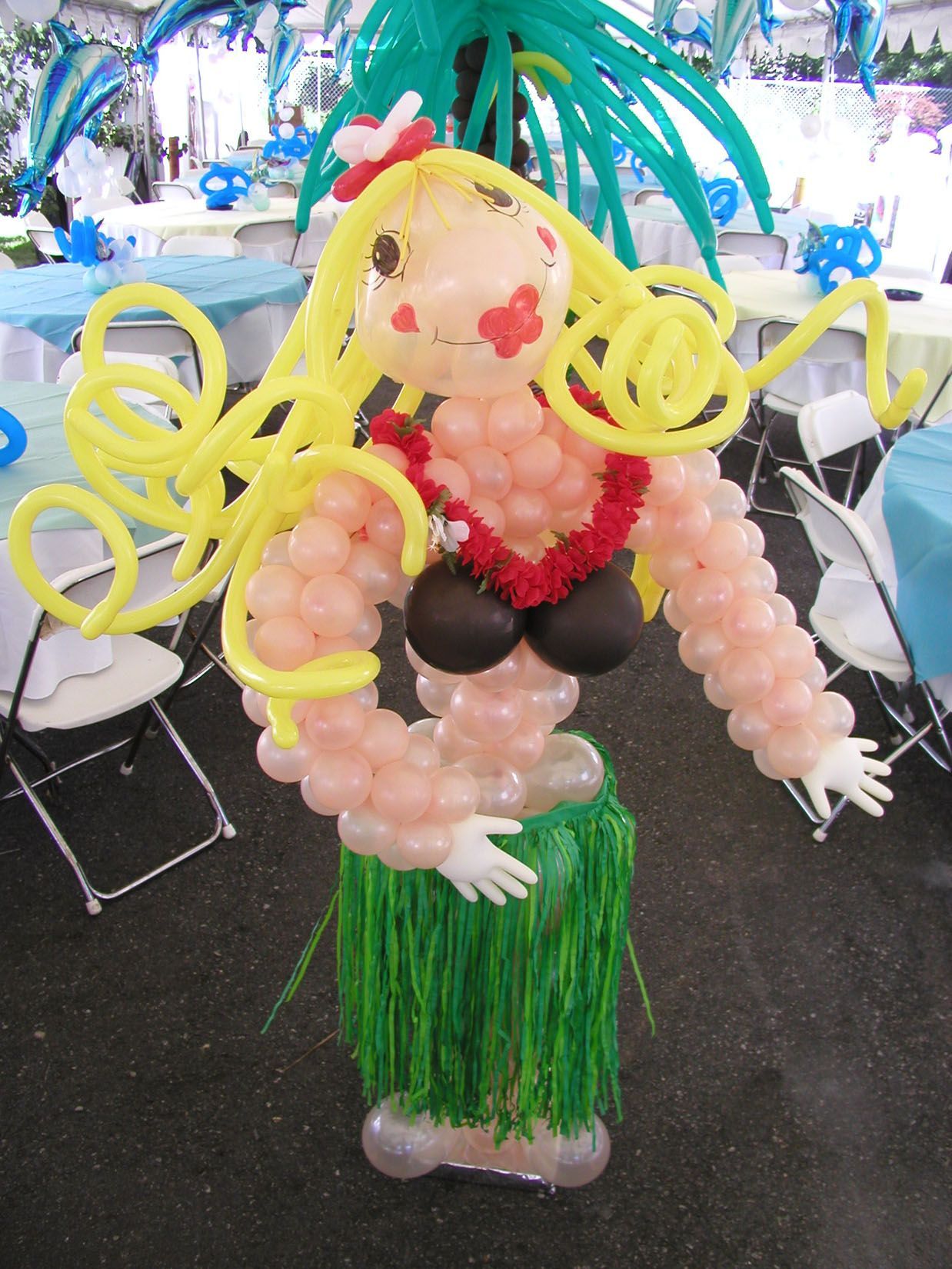 Balloon sculpture of a hula dancer with blonde hair, red flower, lei, and grass skirt, at a party.