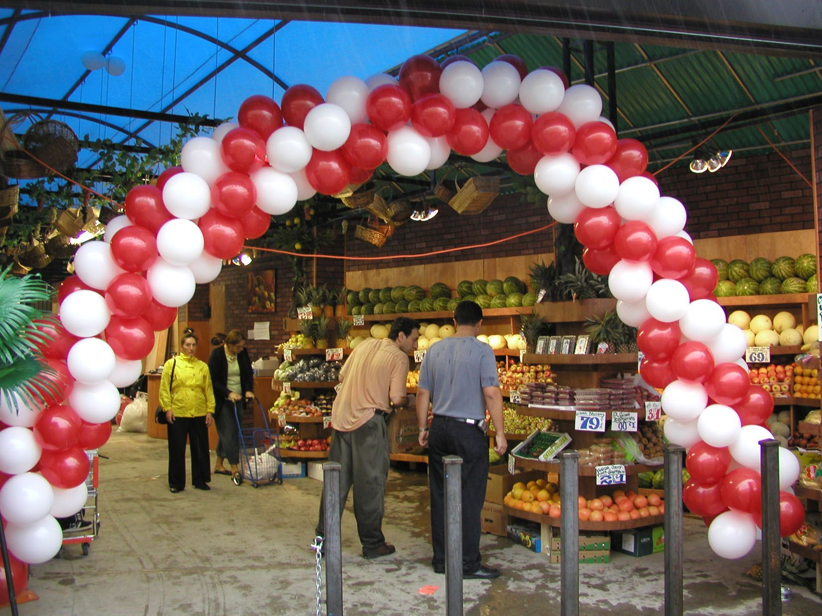 Balloon arch entrance to a produce market, red and white balloons, people browsing.