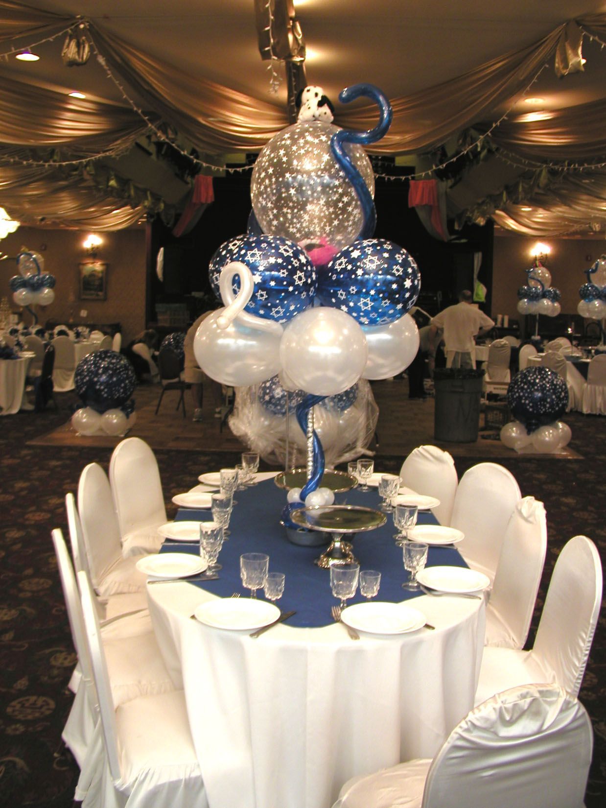 Round table set for a formal event, featuring a blue and silver balloon centerpiece.