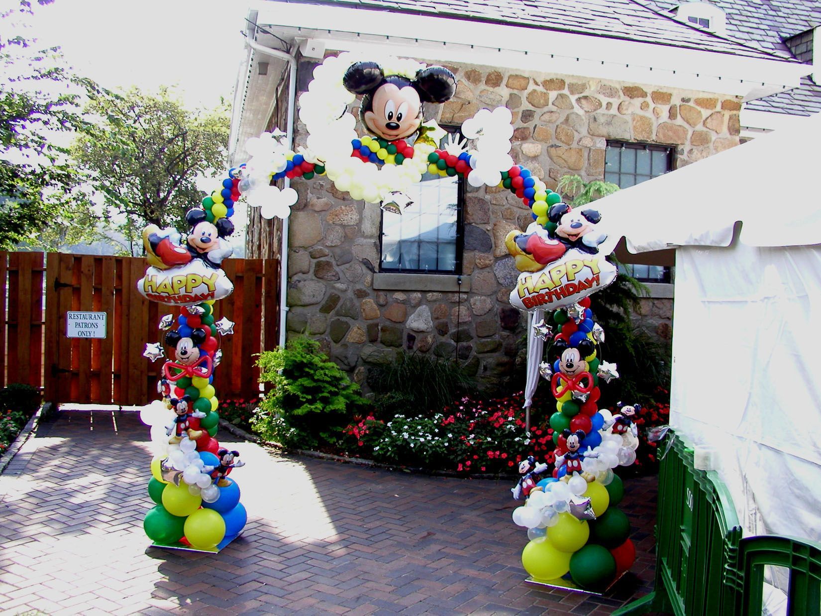 Balloon arch featuring Mickey Mouse at an outdoor event entrance; colorful balloons, stone building background.