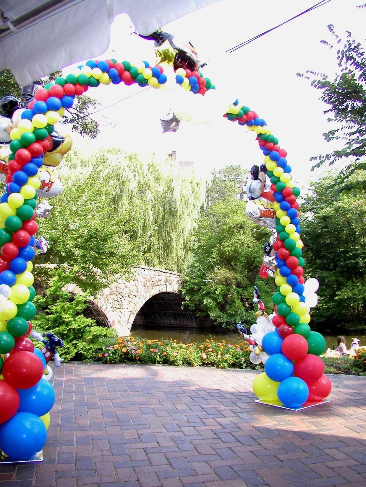Colorful balloon arch over a brick pathway, leading to a stone bridge in a garden.