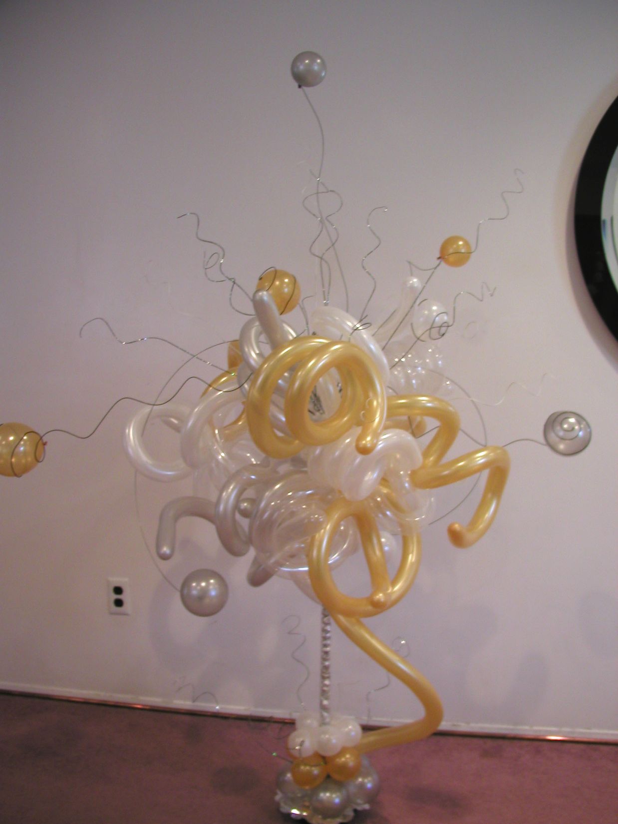 Balloon sculpture of white, gold, and silver orbs and swirls on a silver pole, against a white wall.