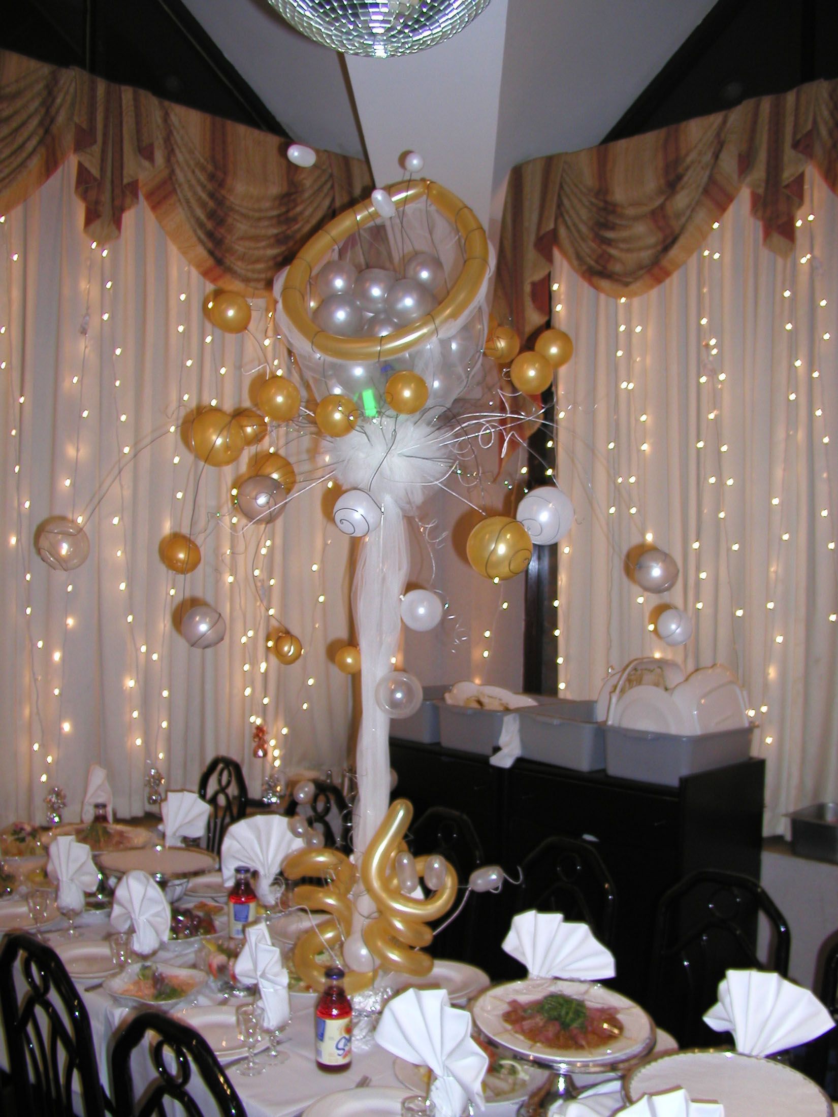 Balloon centerpiece on a decorated table at an event. Gold and white balloons, draped curtains, and string lights.