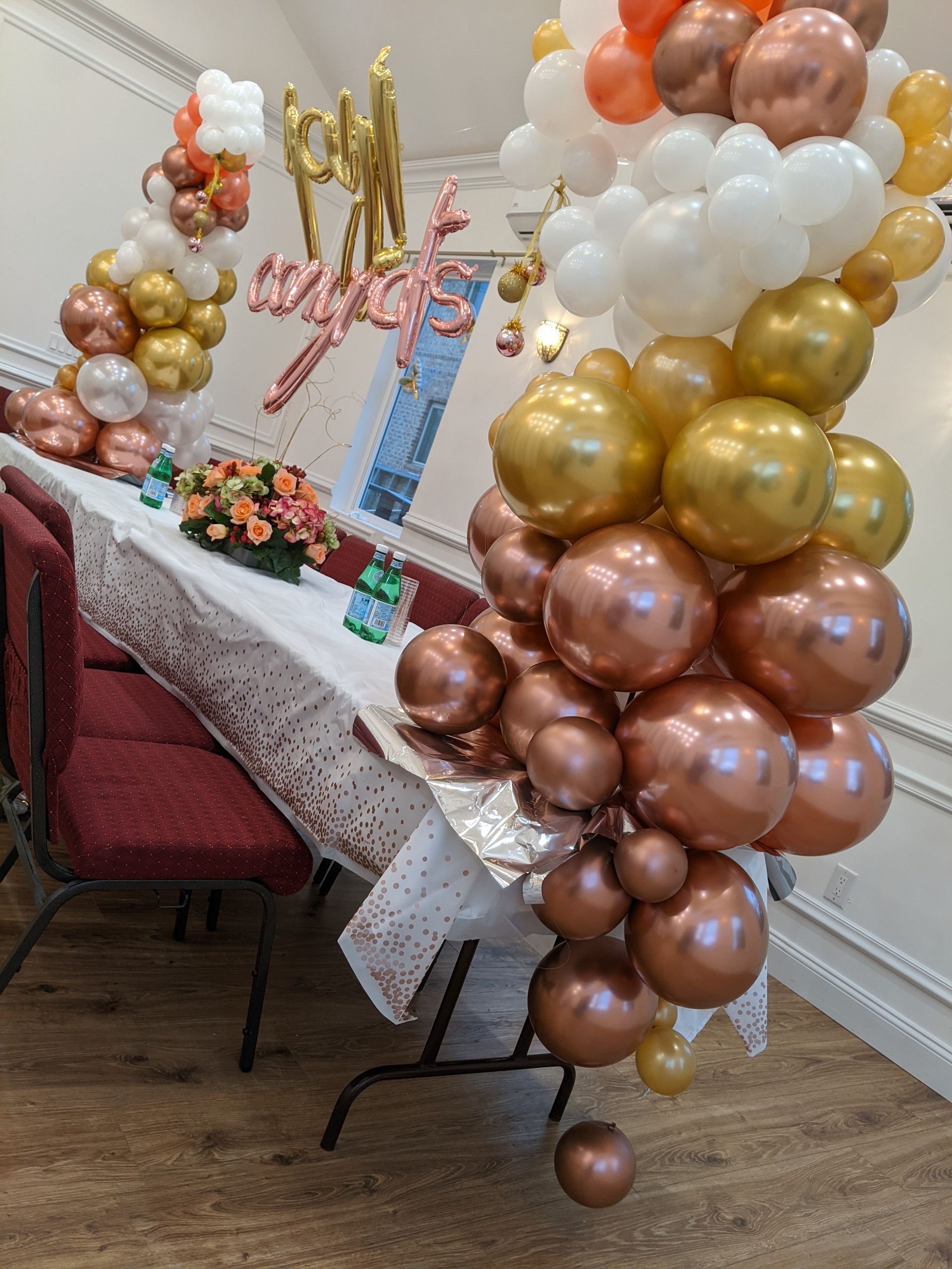 Party decorations: balloon arch in gold, white, copper colors. Table with floral arrangement and 