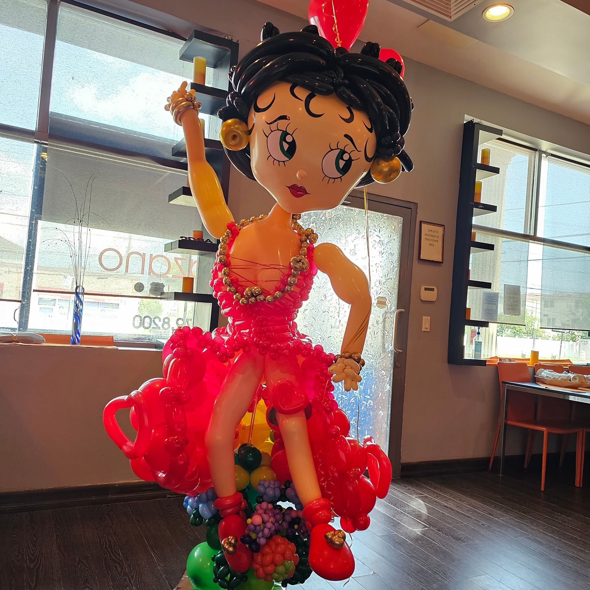 Balloon sculpture of Betty Boop in a red dress, standing on a floral balloon base in a restaurant.
