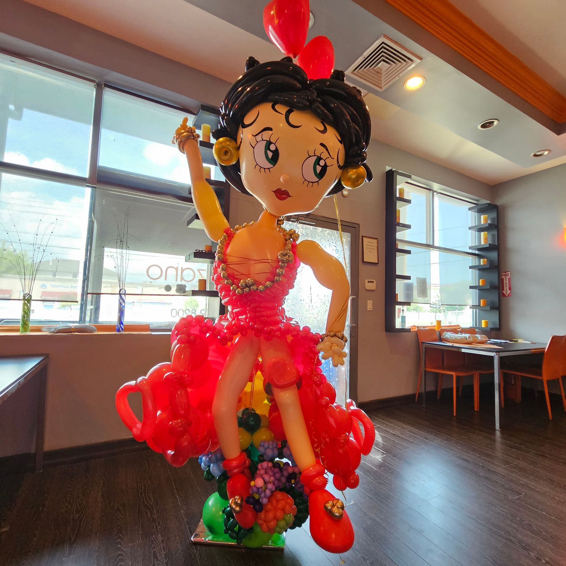 A Betty Boop balloon sculpture with red dress and flowers, inside a restaurant with windows.