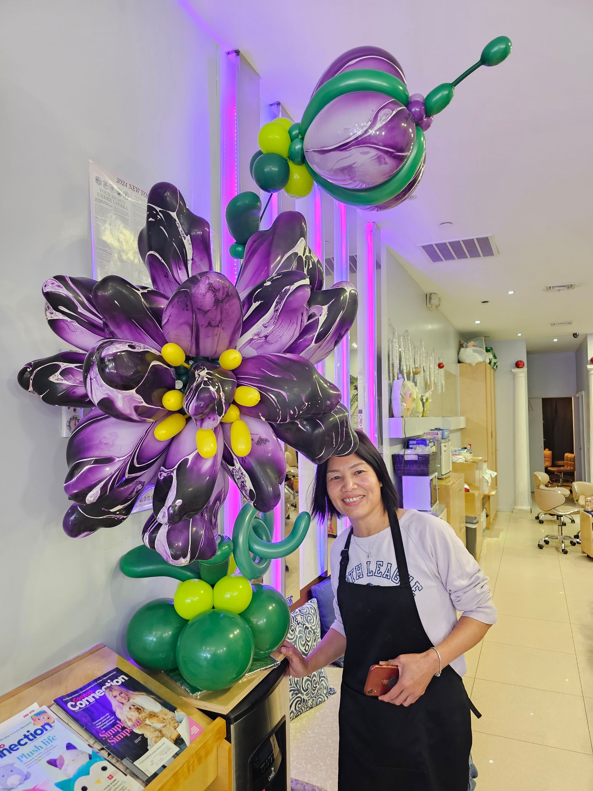 Woman smiles next to a large purple and green balloon flower decoration inside a shop.