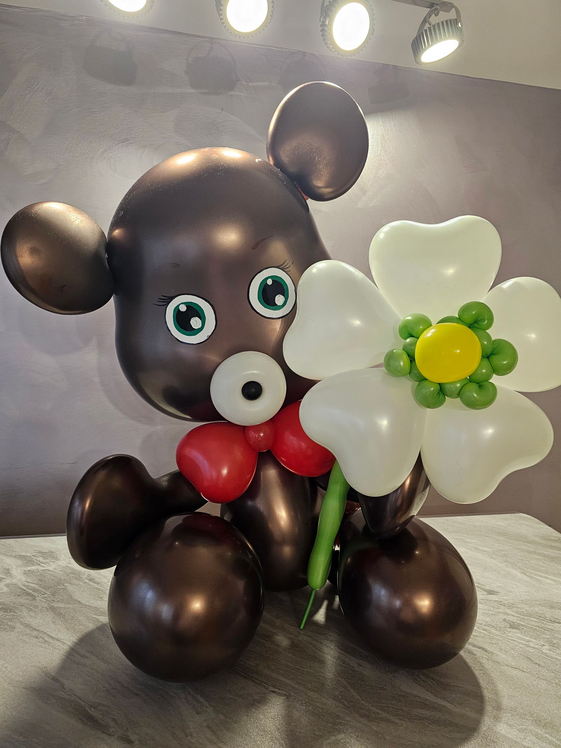Brown balloon bear holding a white flower; sitting on a surface.