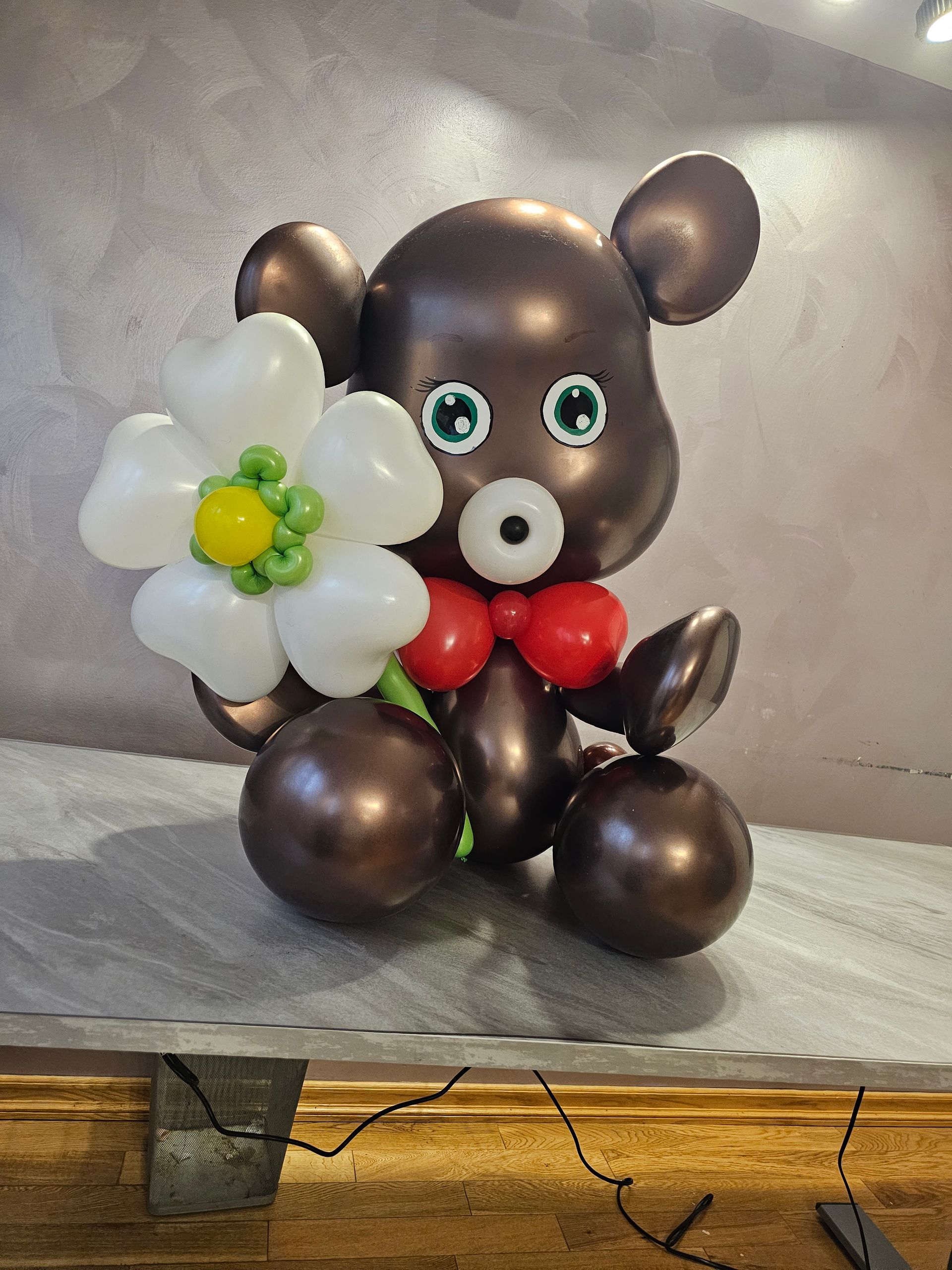 Brown balloon bear holding a white balloon flower with green and yellow accents, red bow tie.