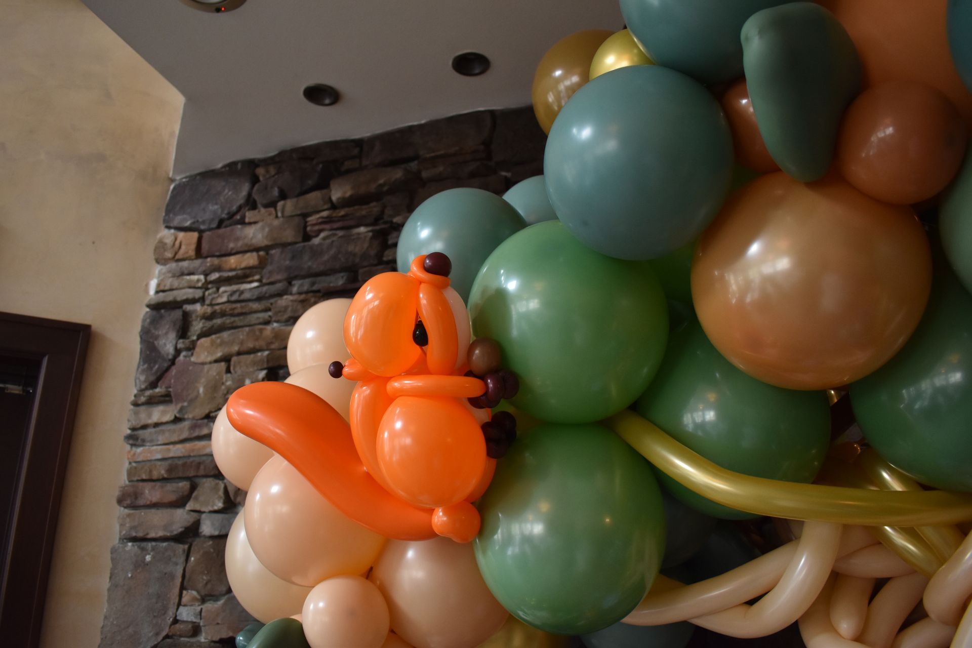 Balloon squirrel climbing a cluster of colorful balloons, set against a stone wall.