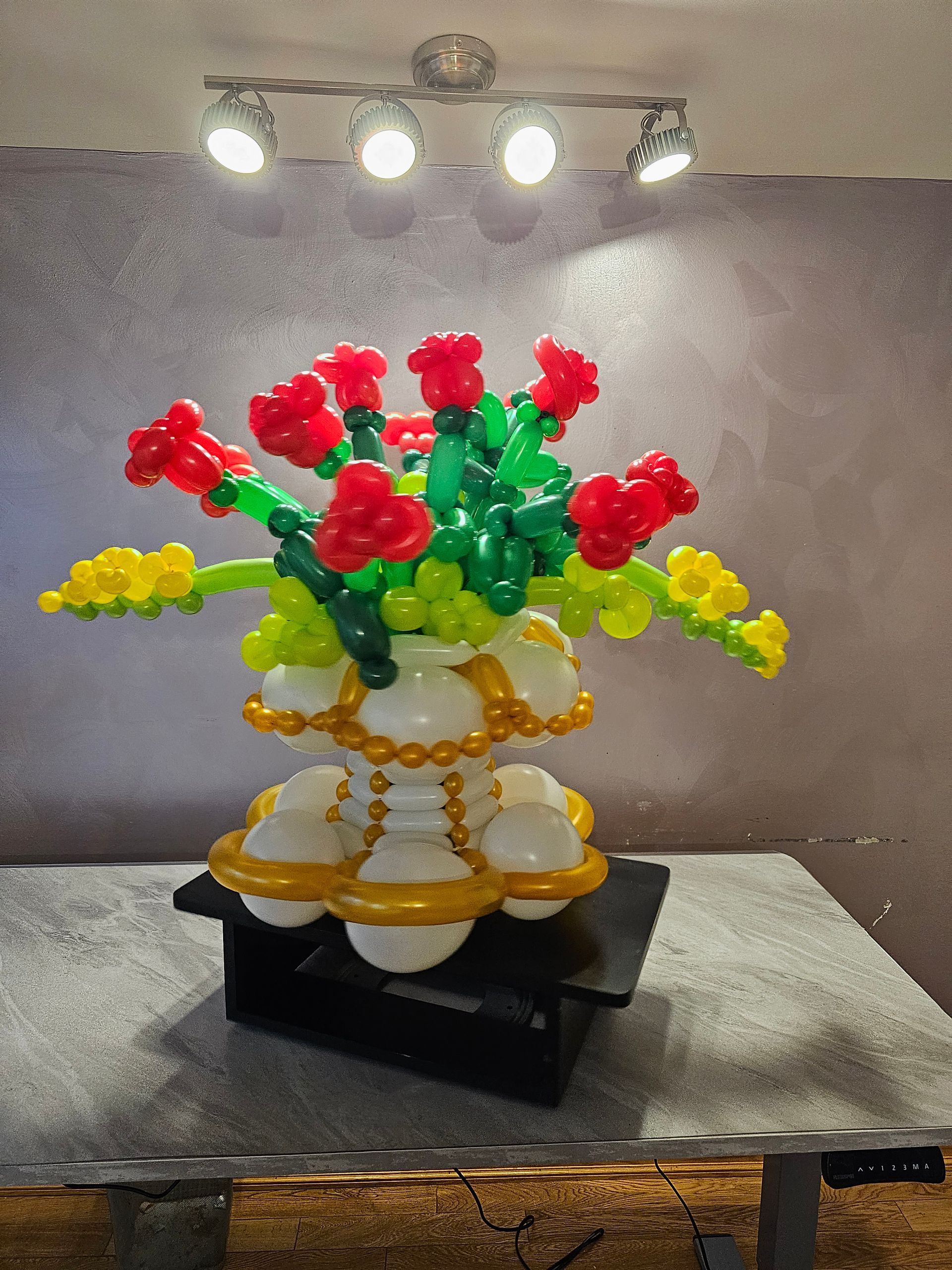Balloon sculpture of red and yellow flowers in a white and gold vase on a black stand.
