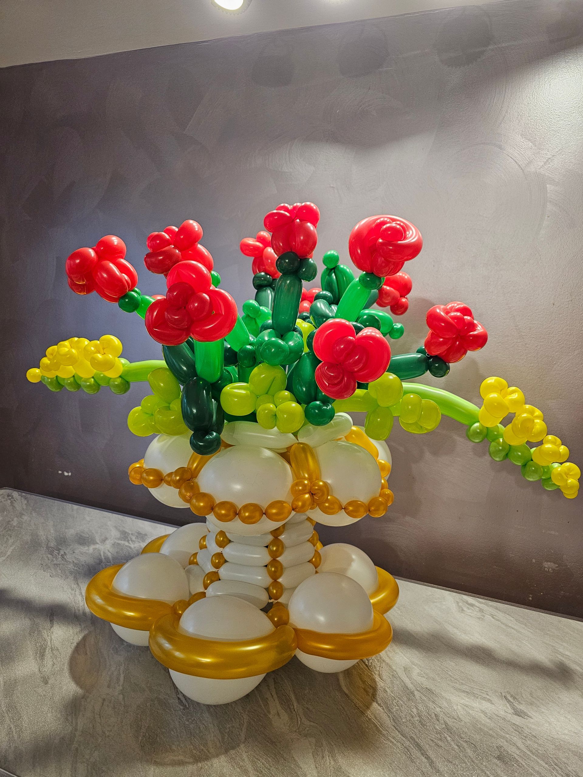 Balloon sculpture of red roses and greenery in a white and gold vase.