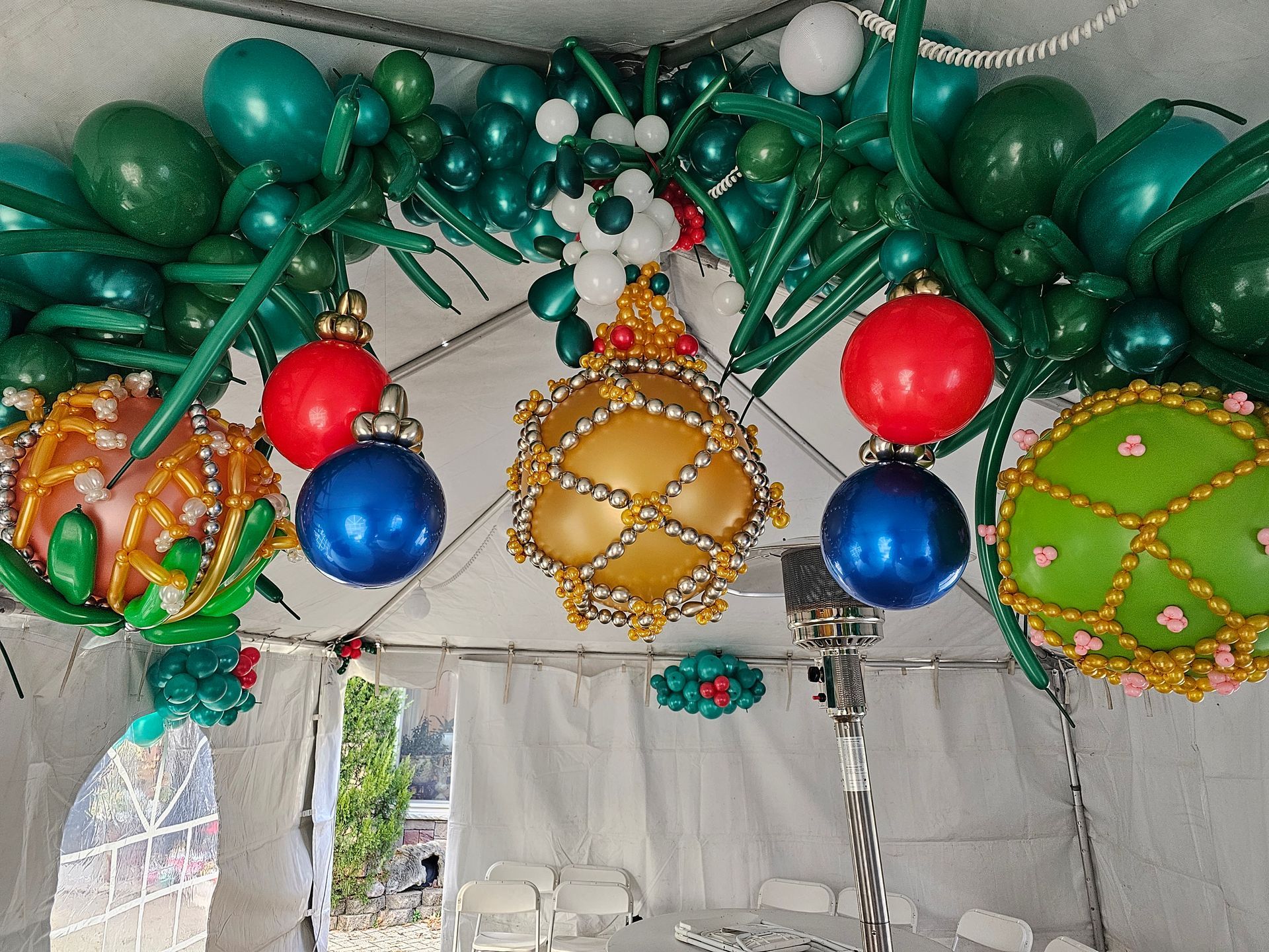 Festive balloon arch with red, blue, and gold ornaments, greenery, and white tent background.