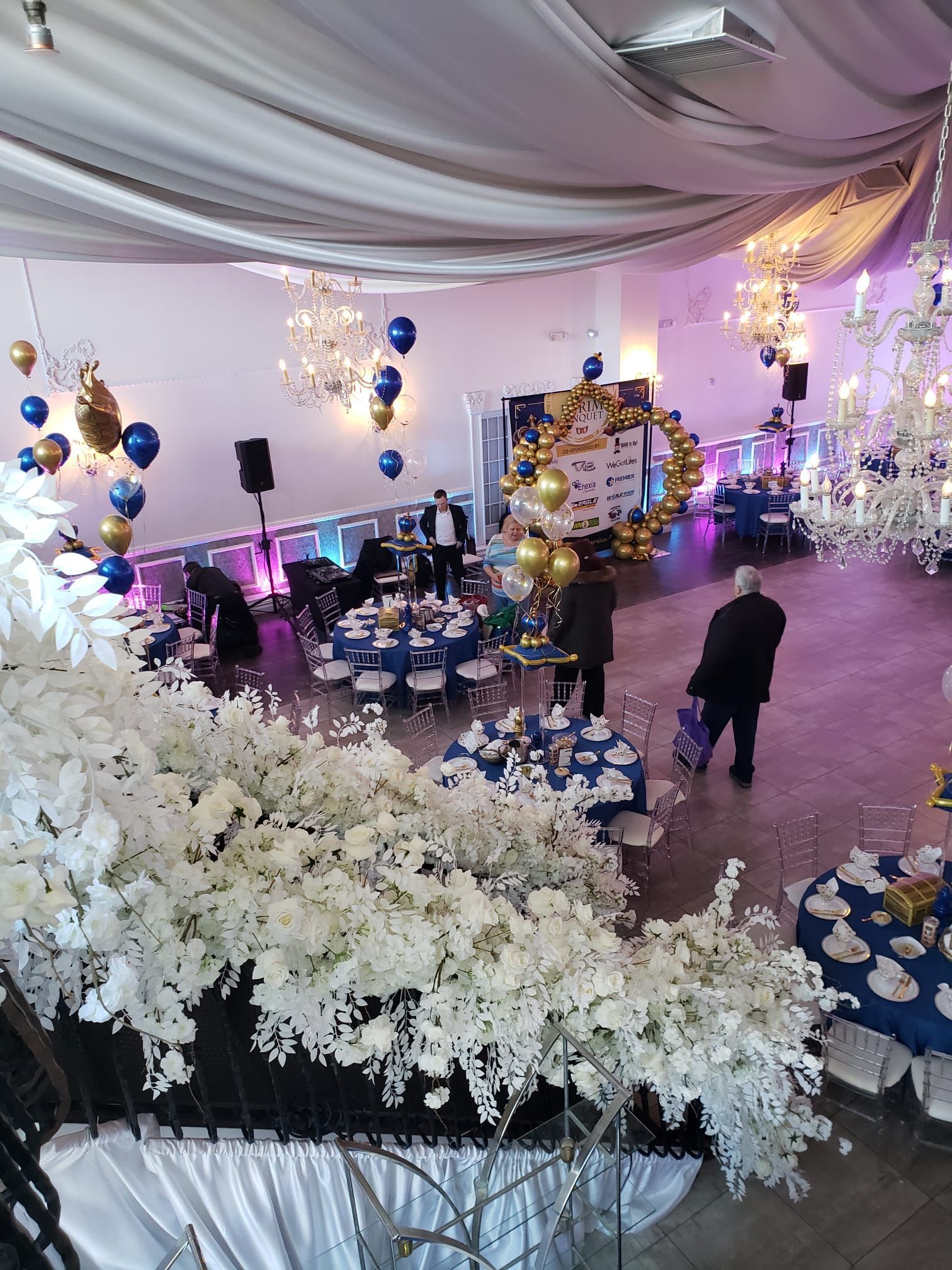 Elegant event venue with tables, floral arrangements, chandeliers, and balloons. People are setting up.