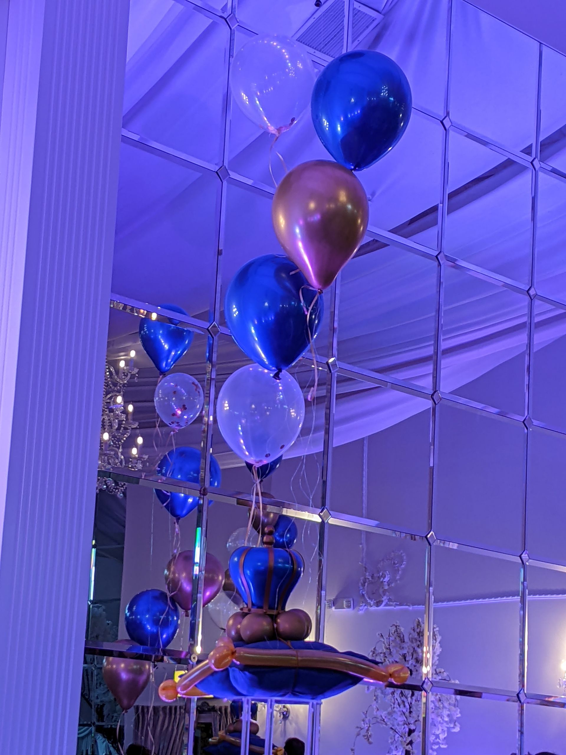 Balloons in various colors floating against a mirrored wall. Includes blue, gold, and clear balloons.