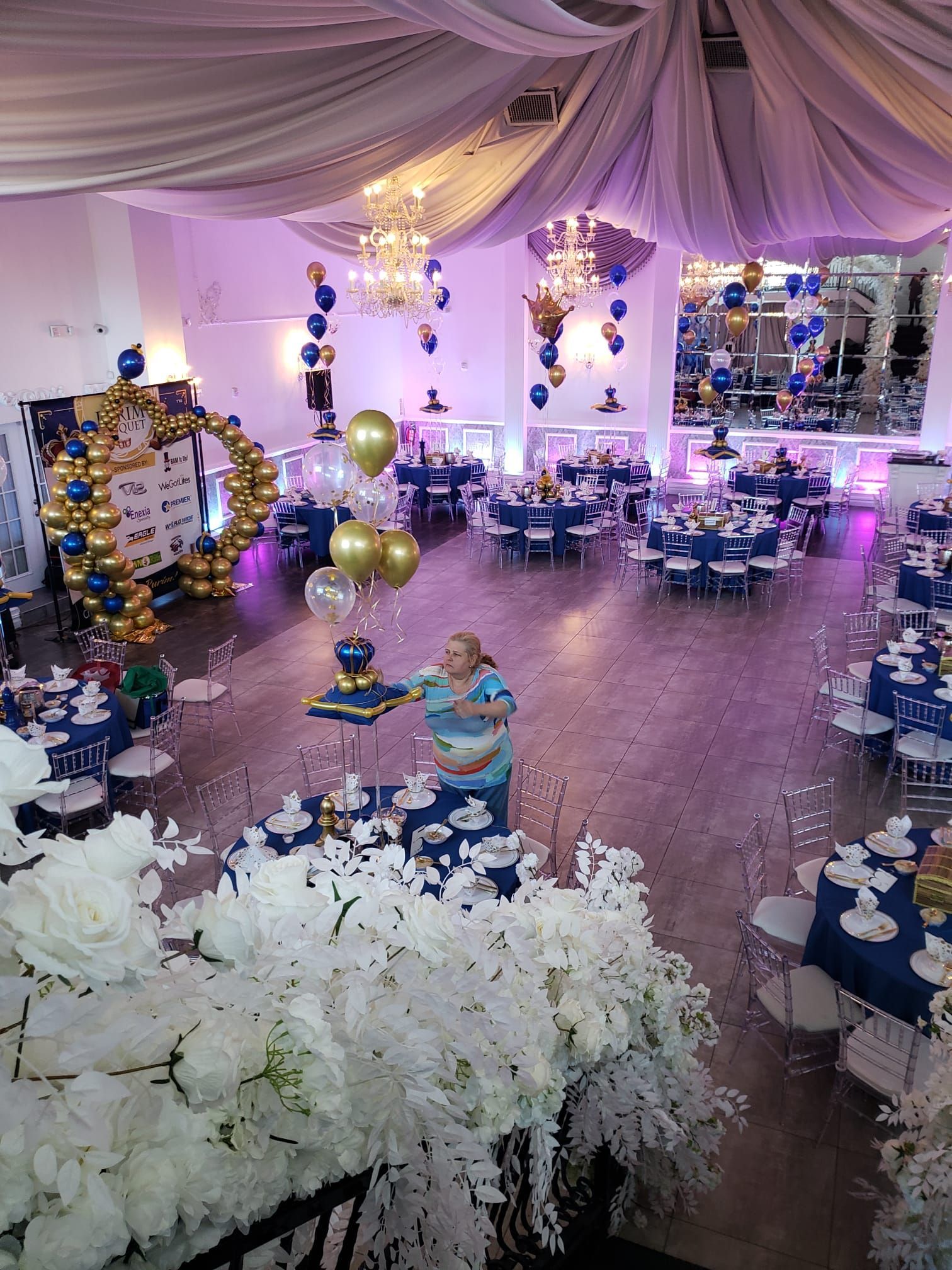 Event hall decorated in blue and gold. Woman arranging items at a decorated table, guests seated at tables.