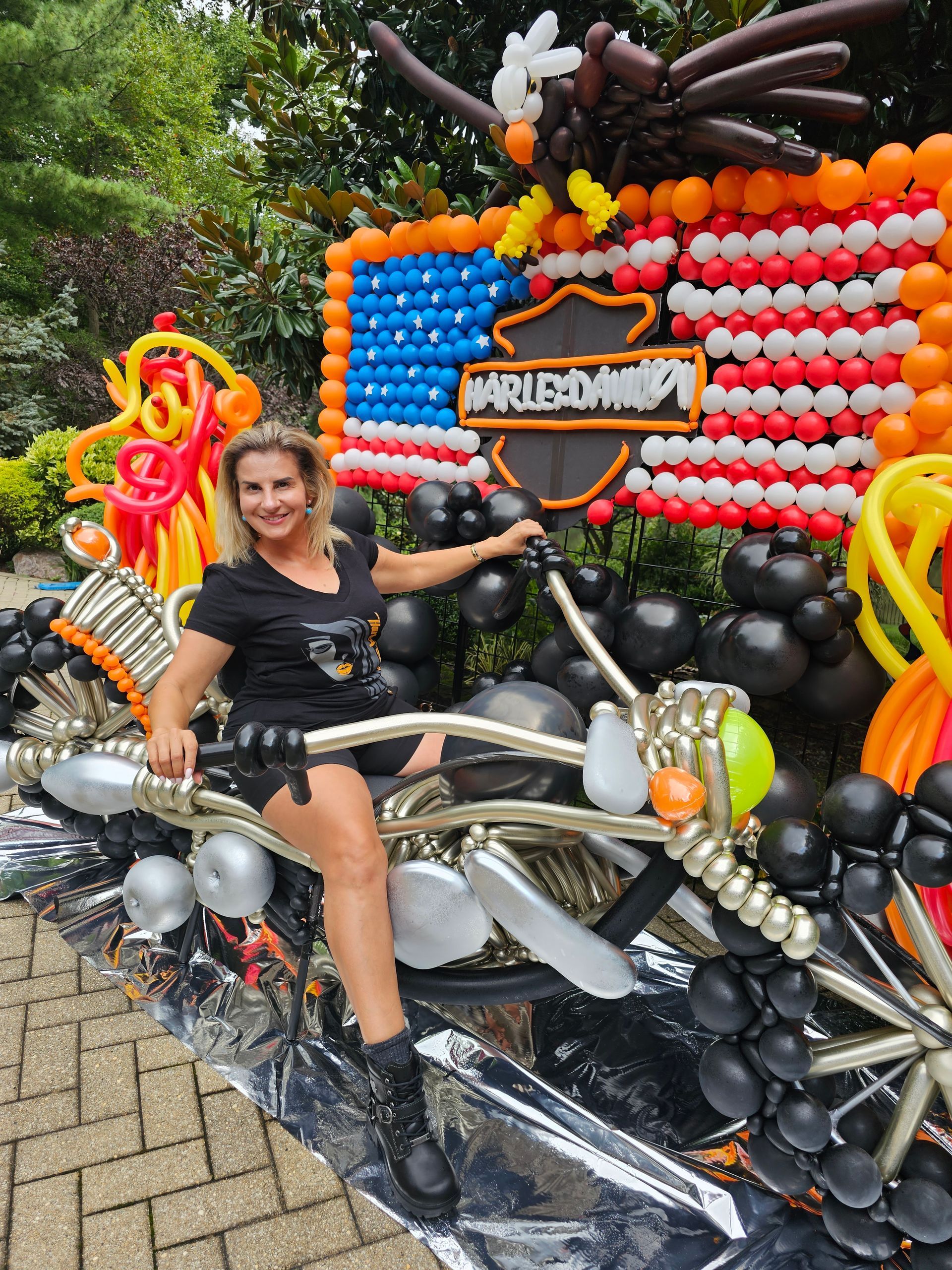 Woman posing on a balloon motorcycle sculpture with a Harley-Davidson flag background.