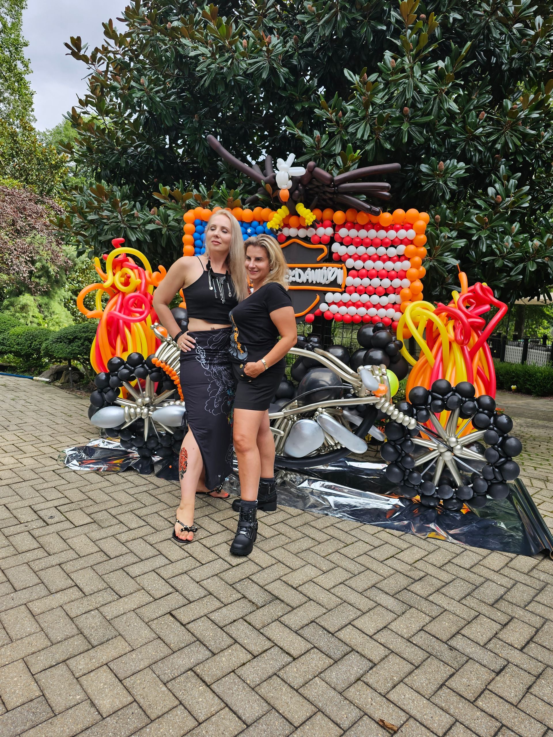 Two women pose with a balloon motorcycle sculpture, a large eagle on top. Outdoors, brick patio.