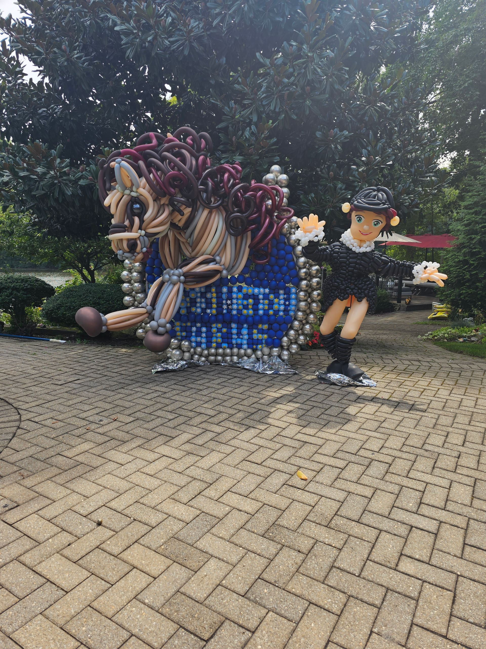 Sculpture of a boy pulling a horse-drawn carriage made of bottle caps on a brick path, trees in the background.