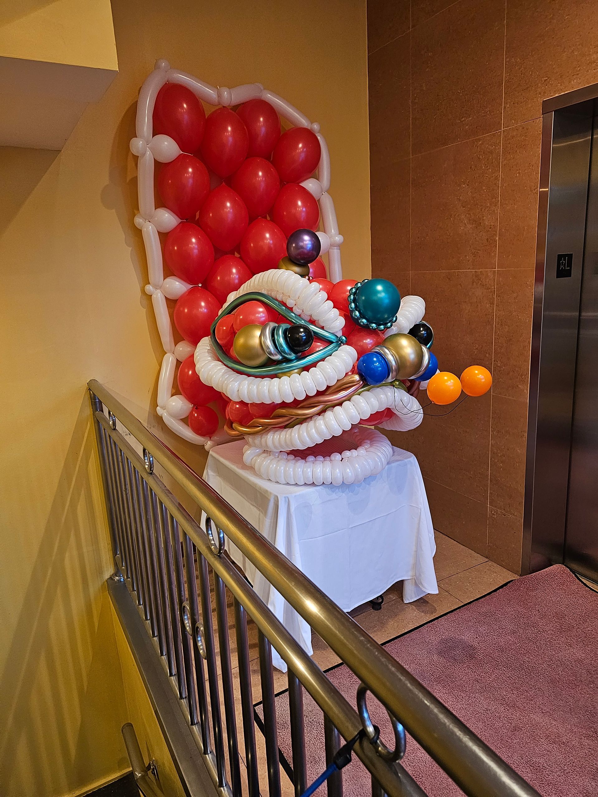 Balloon lion head sculpture with a red balloon throne, near stairs and elevator.