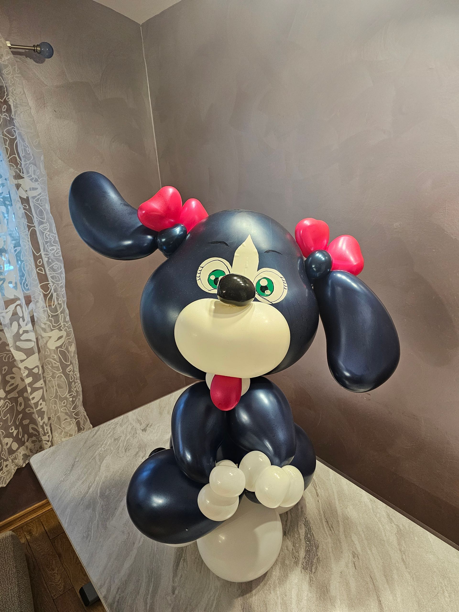 Balloon sculpture of a dark blue dog with white face, pink bows, and green eyes sitting on a white balloon base.
