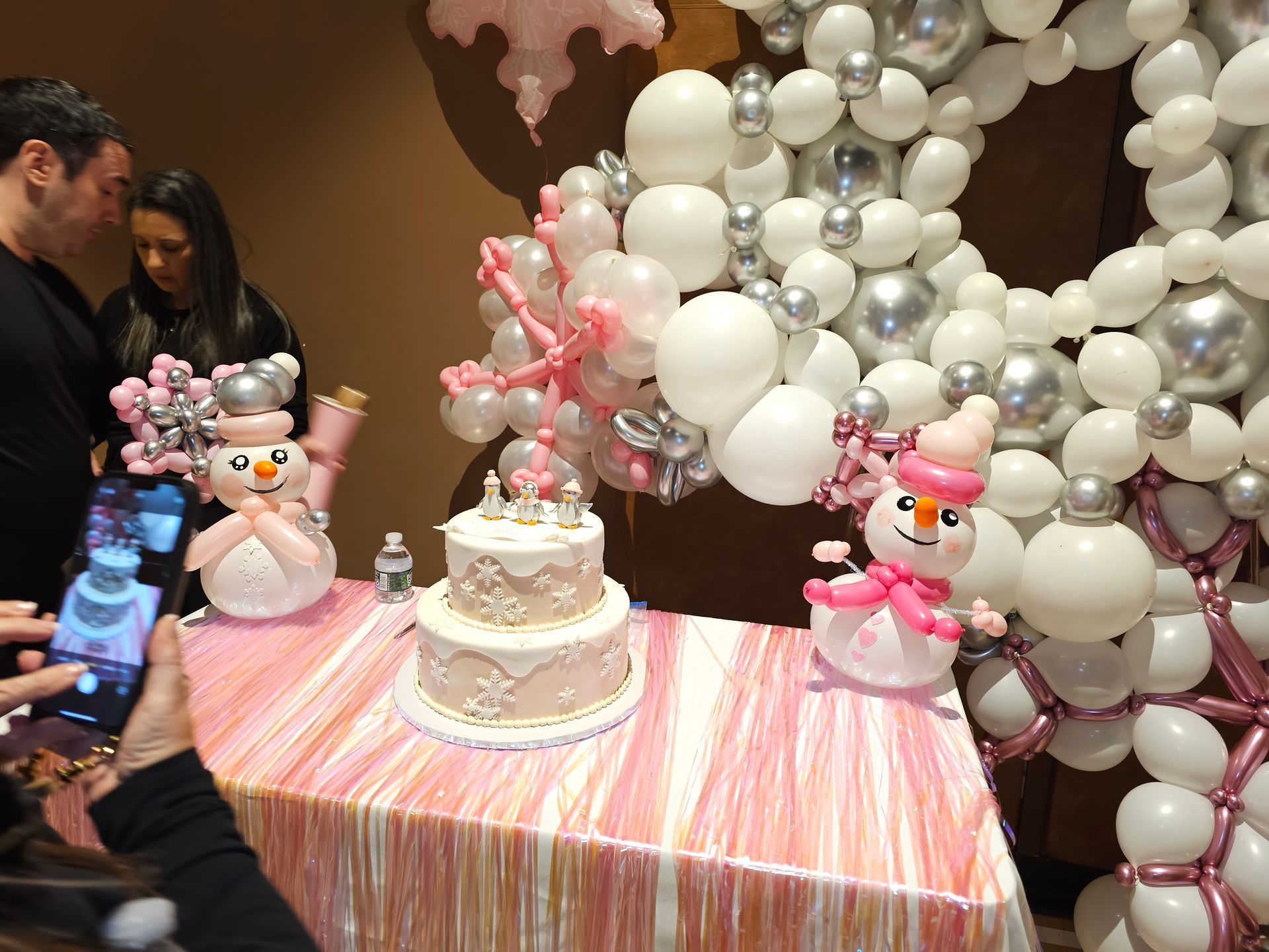 Snow-themed birthday party: cake, balloon snowman, silver & white balloons, and people around a table.