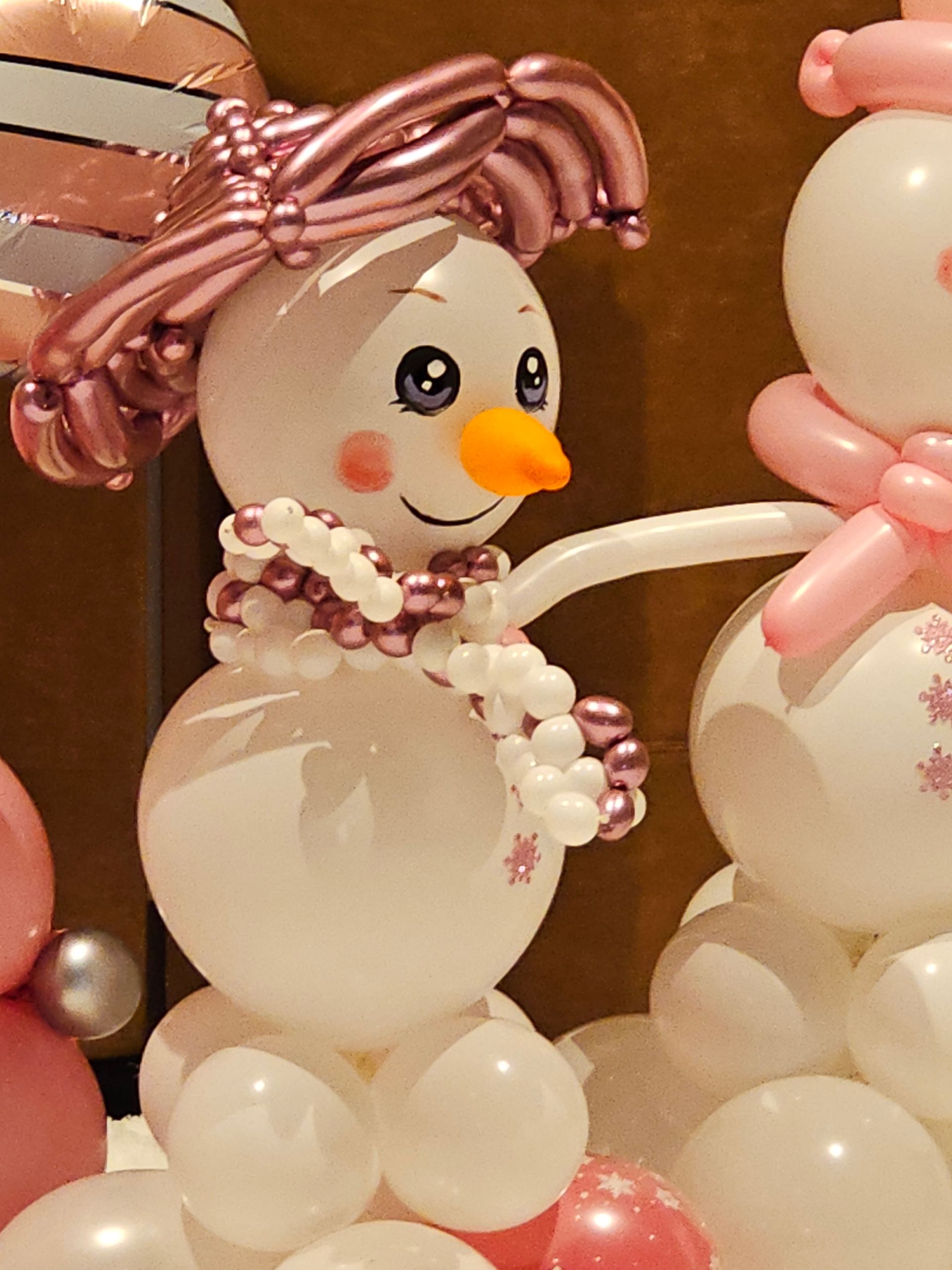 Balloon snowman with pink scarf, hat, and arms, smiling with orange nose.