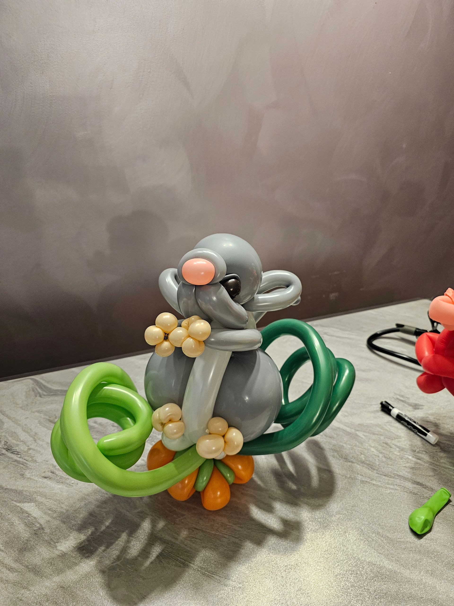 Gray balloon mouse sculpture with green tail, orange feet, and tan flowers.