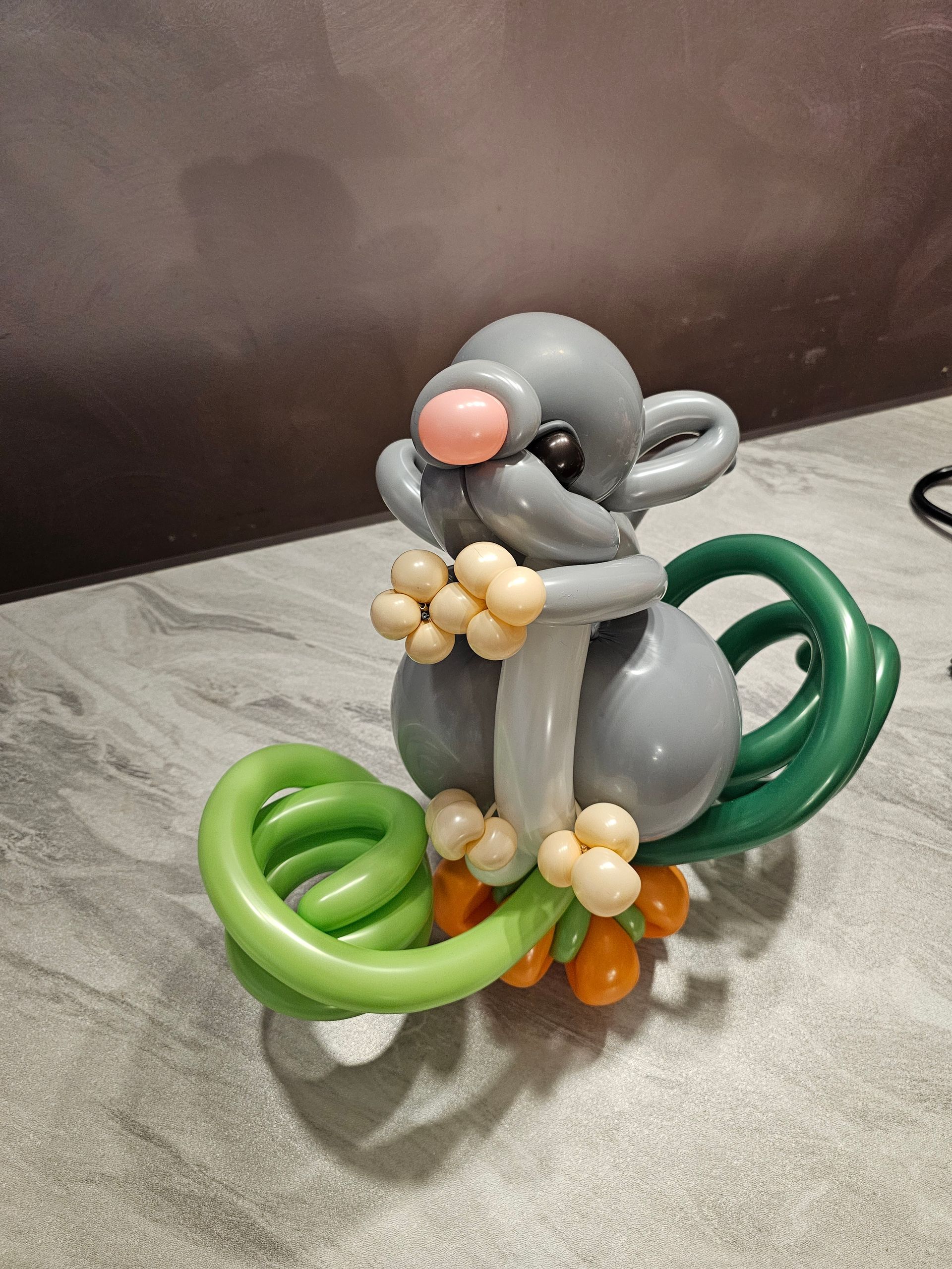 Balloon sculpture of a gray mouse with a pink nose, holding yellow fruit, and a green tail.