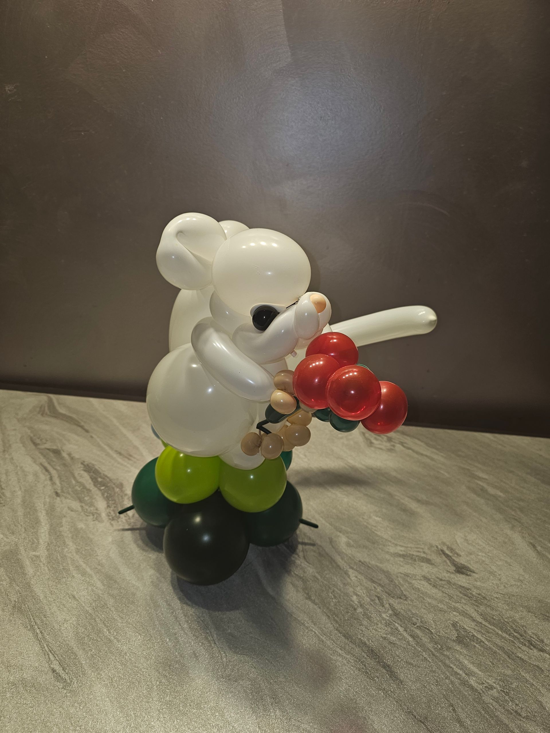 Balloon sculpture of a white mouse holding red cherry balloons. It stands on green and black balloon base.